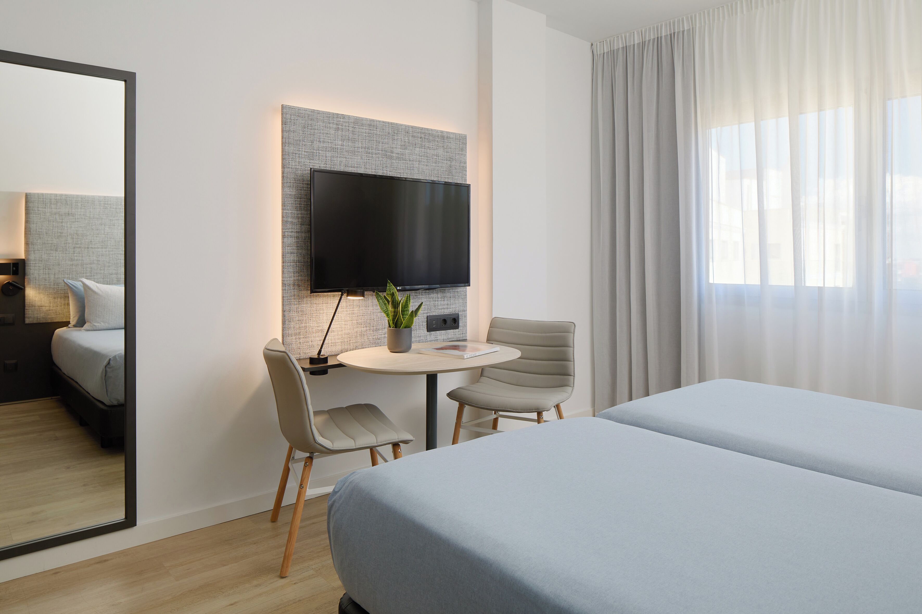 Ett modernt hotellrum med en stor säng, ett skrivbord med en stol, en platt-TV och en stor spegel.