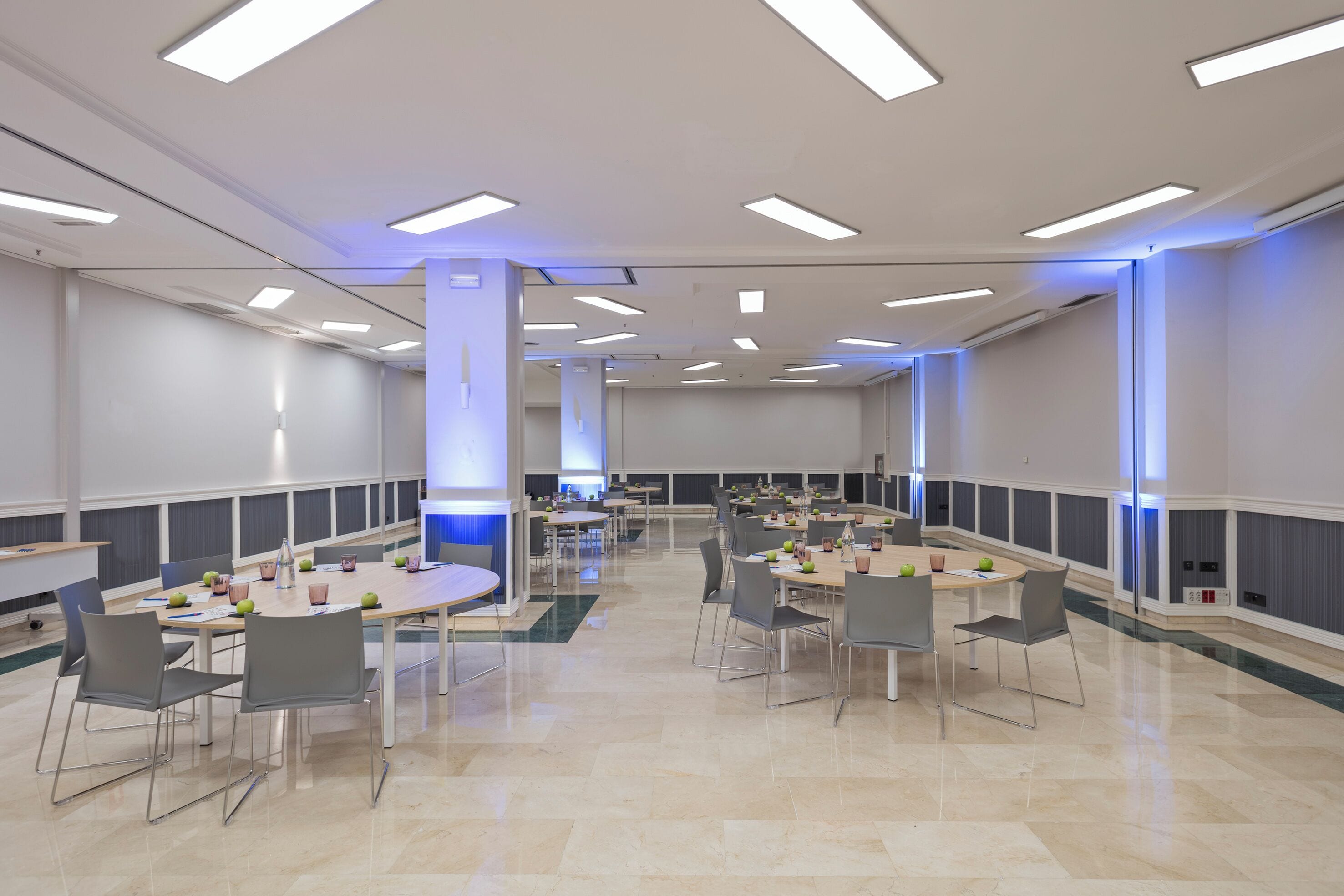 Una amplia sala de conferencias con múltiples mesas y sillas, equipada con iluminación moderna y un diseño limpio y minimalista.