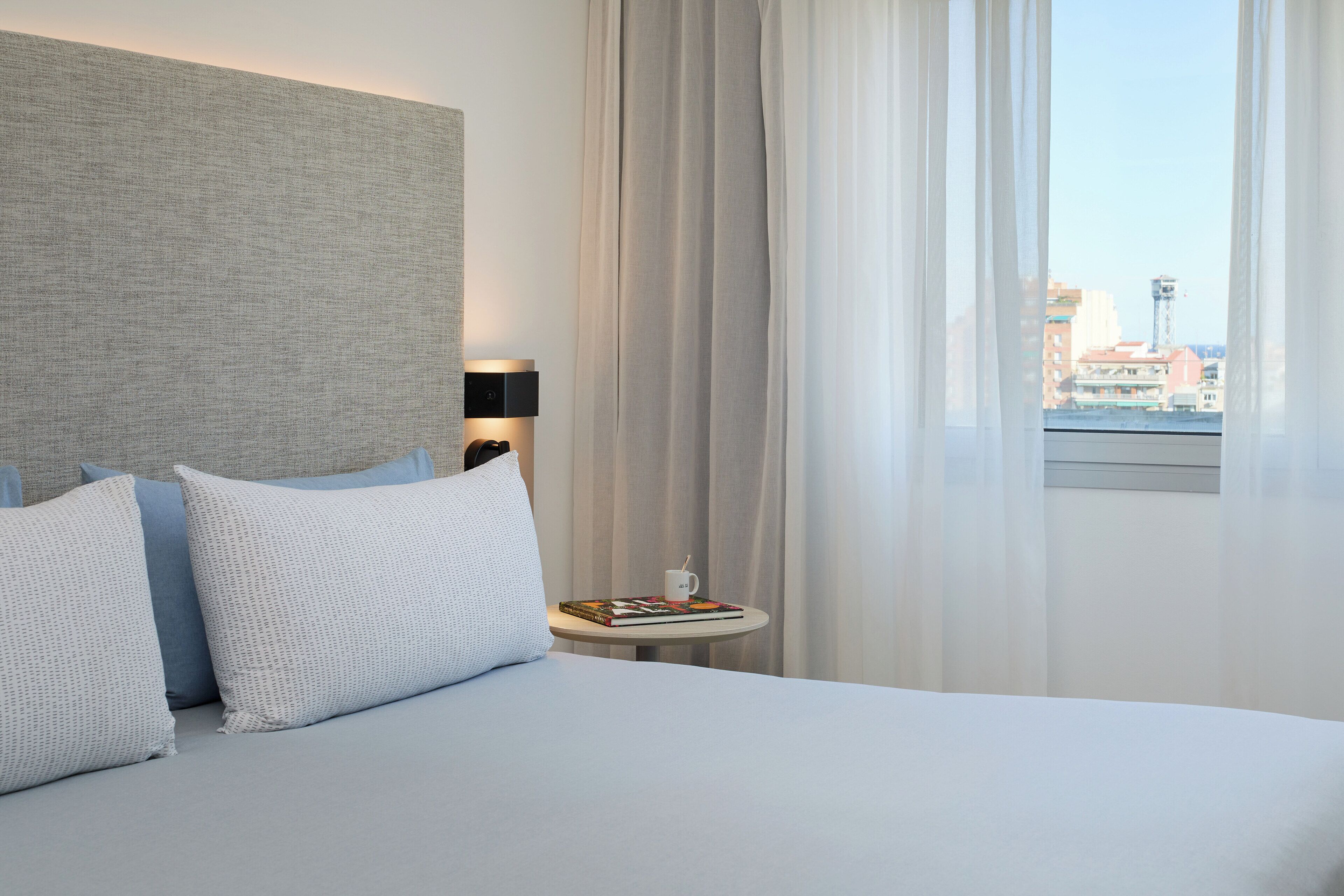 Ein Hotelzimmer mit Bett, Nachttisch, Lampe, einem Fenster mit Vorhängen und Blick auf Gebäude.