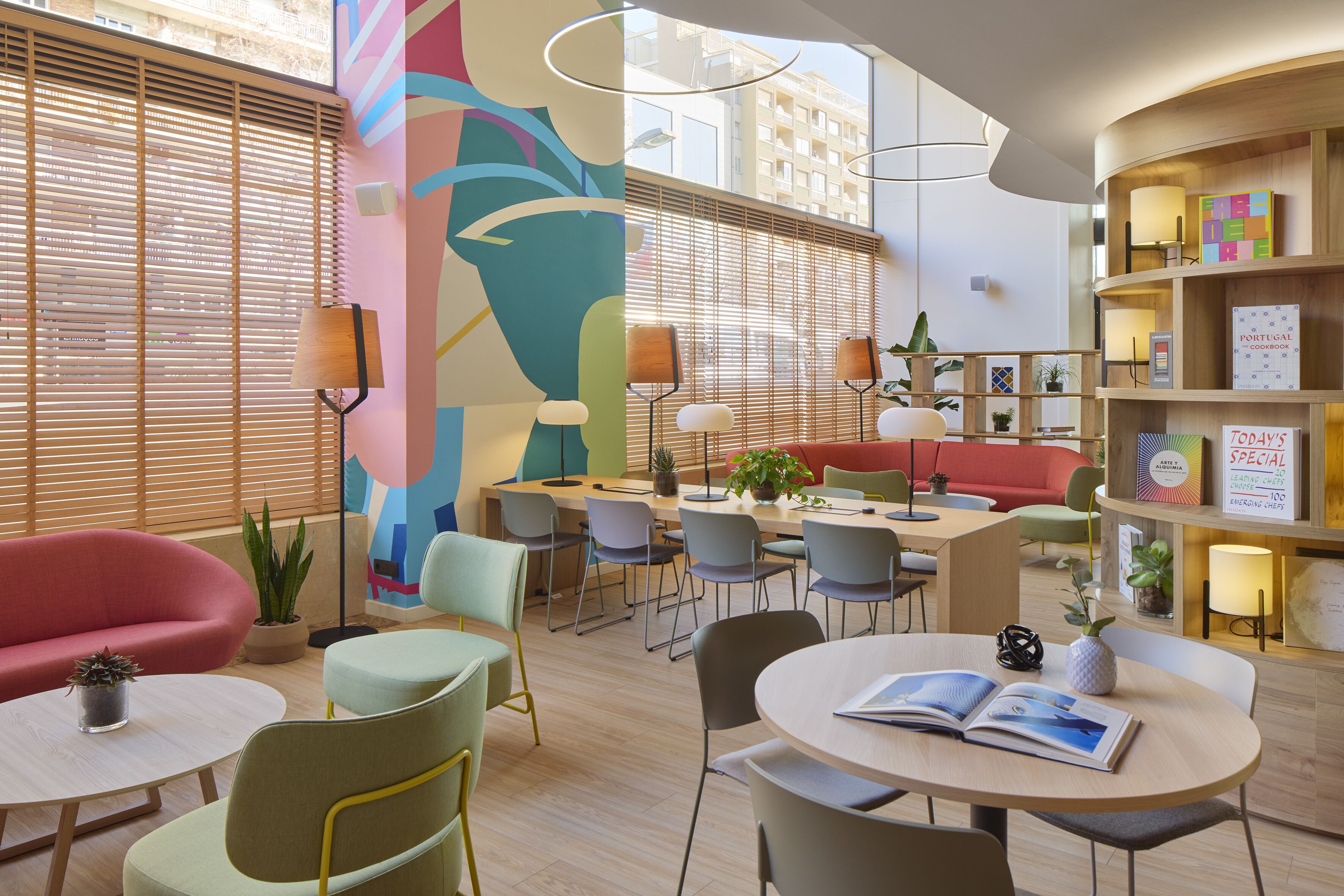 Un moderno vestíbulo de hotel con un área de comedor central, asientos cómodos y un mural vibrante.