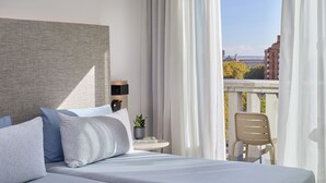 Habitación de hotel con una cama, una mesita con una planta y vistas de la ciudad a través de cortinas translúcidas.
