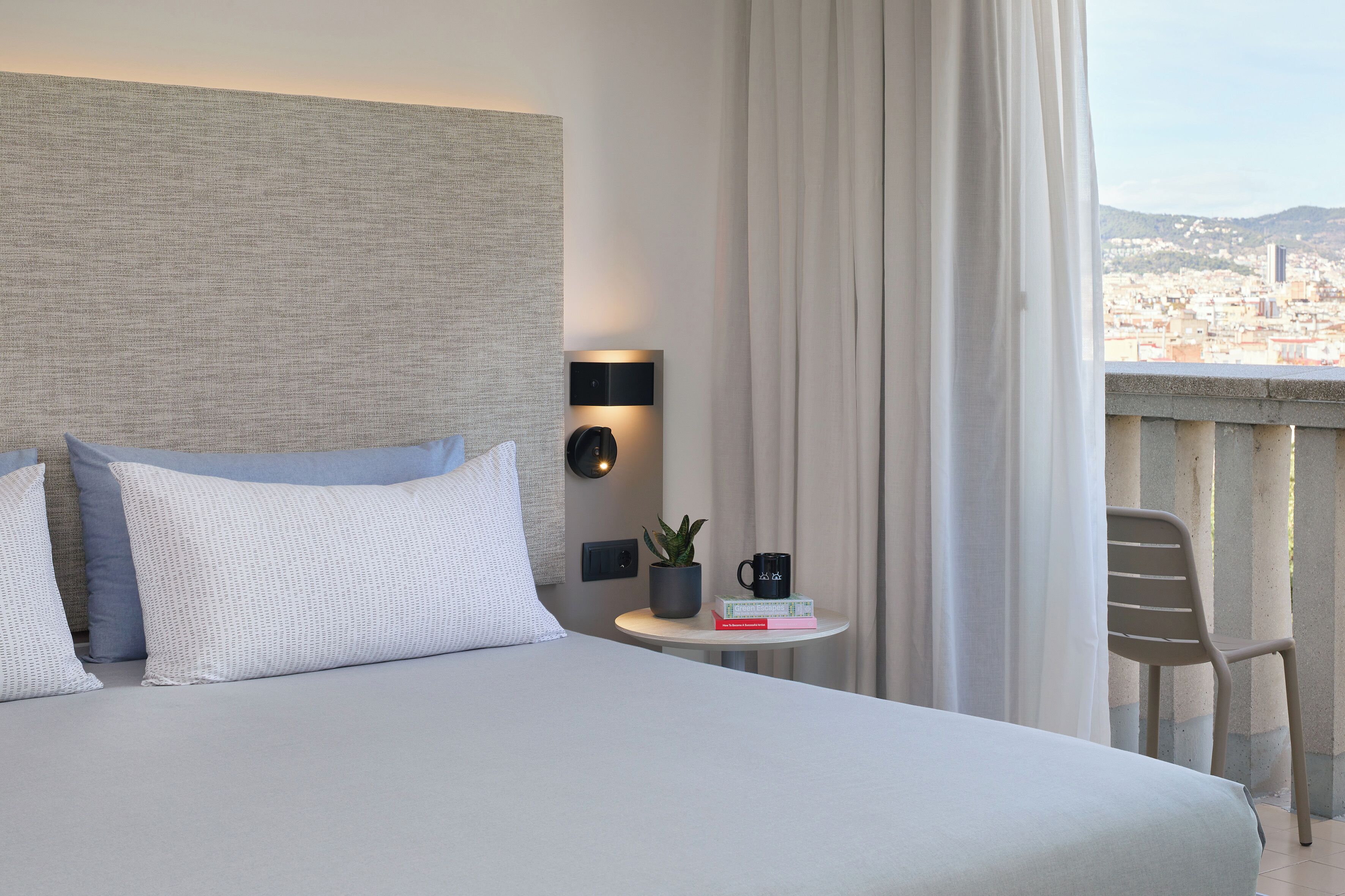 A modern hotel room with a large bed, a small table with a plant and a mug, a chair, and a view of the city.