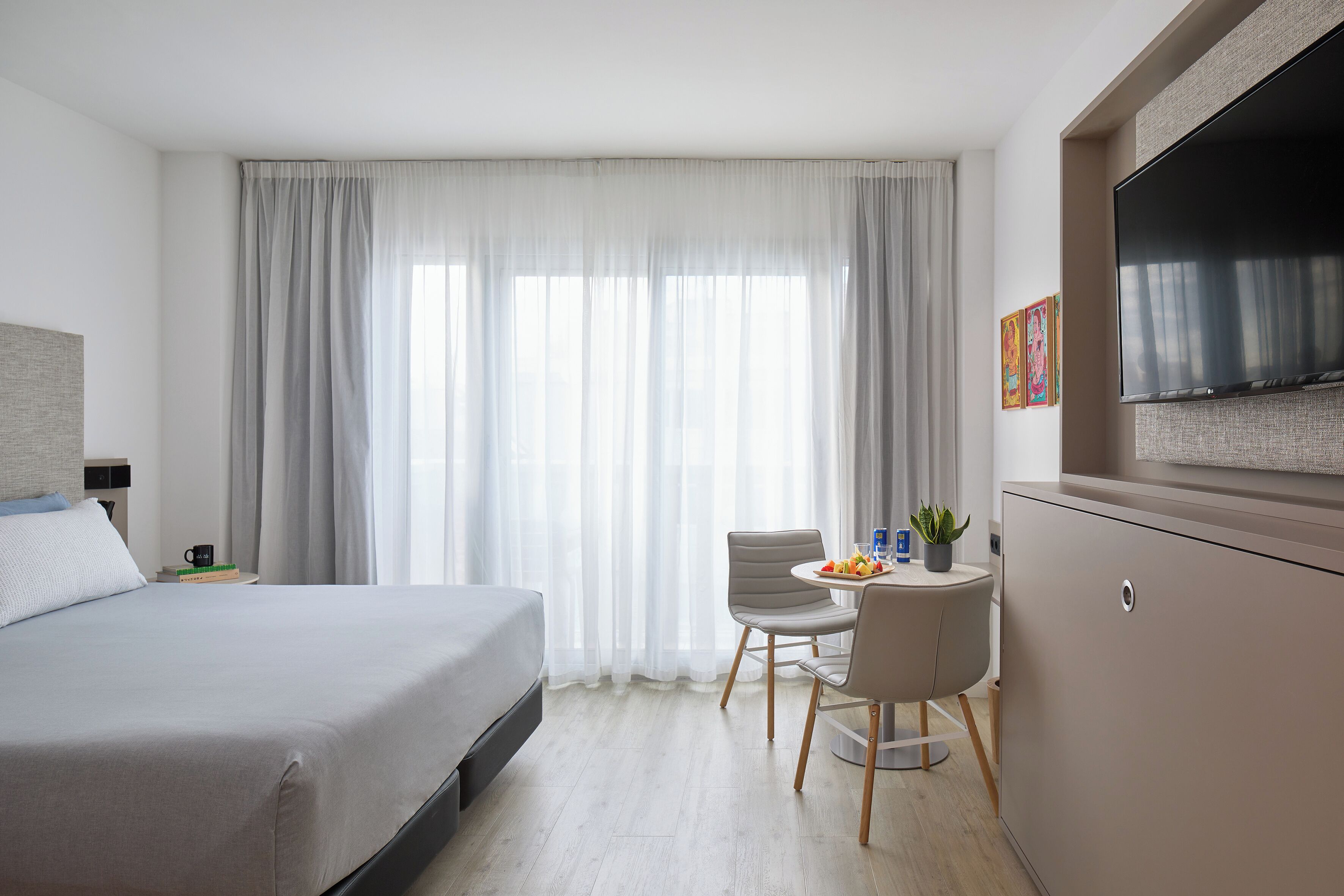 A modern hotel room with a bed, a desk with a chair, a TV, and a large window with curtains.