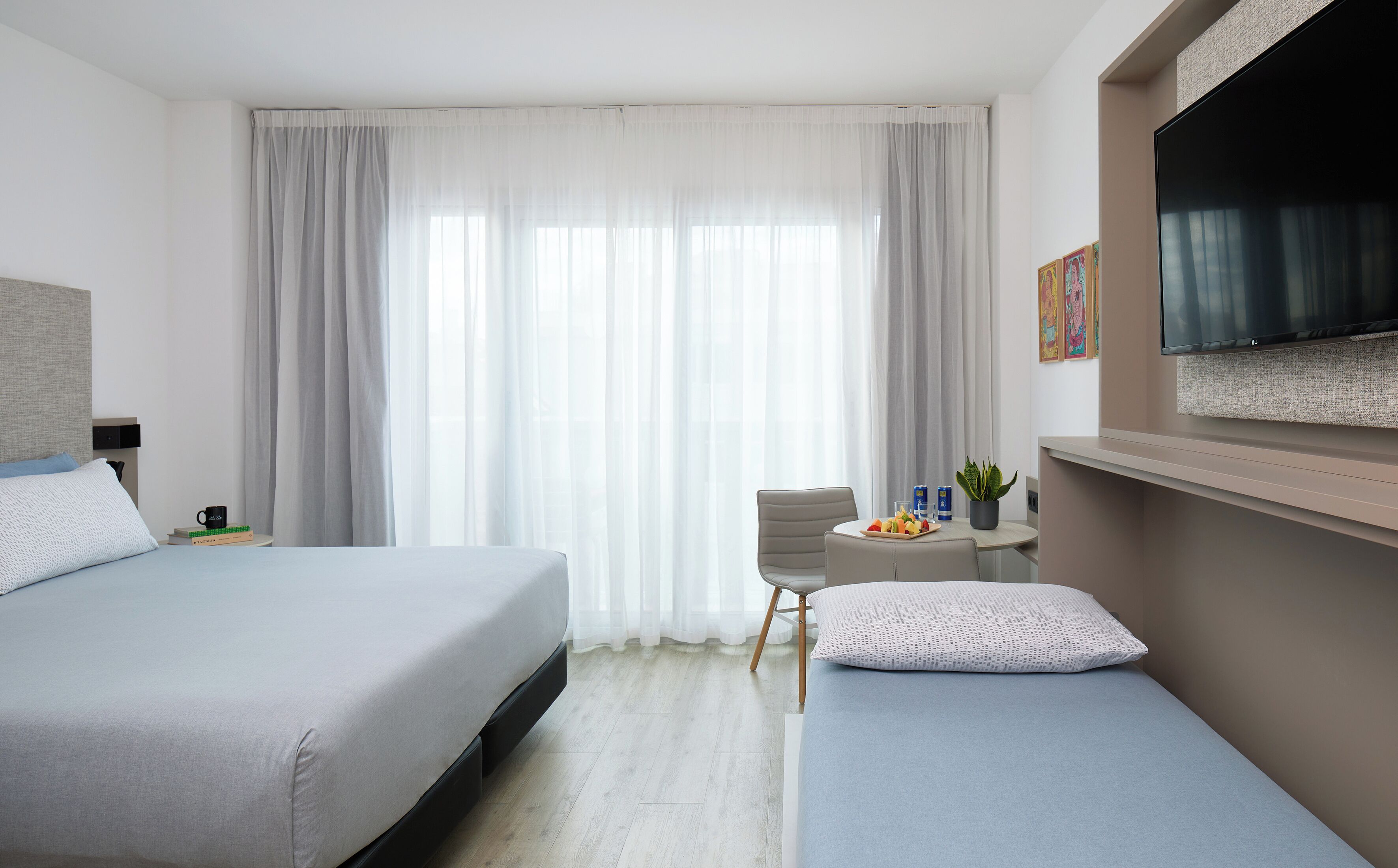 A hotel room with a bed, a chair, a small table with a tray of fruit, a plant, and a TV mounted on the wall.