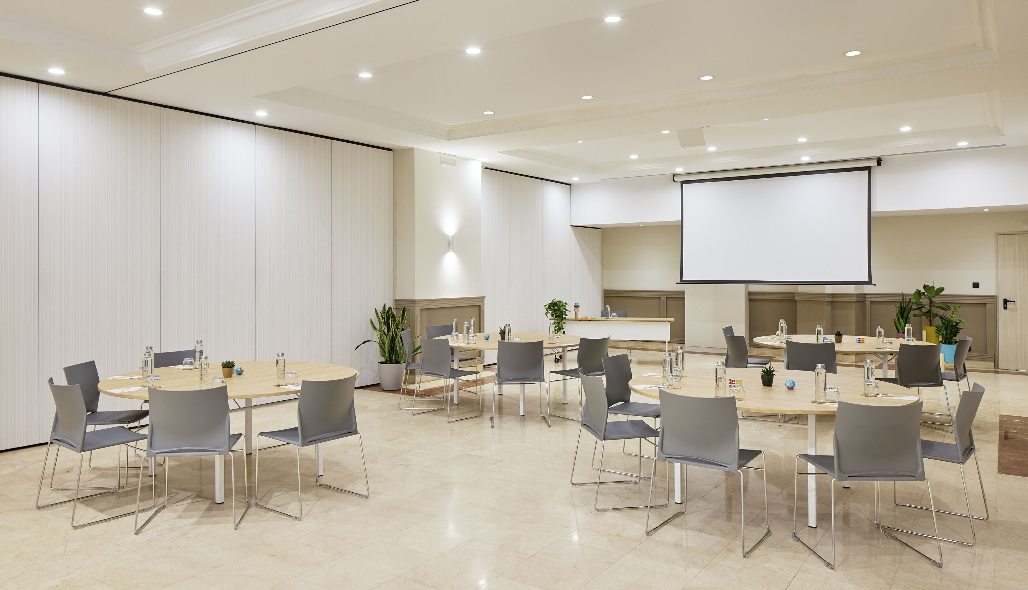 Un salón de conferencias con mesas redondas, sillas grises y una pantalla grande para presentaciones.