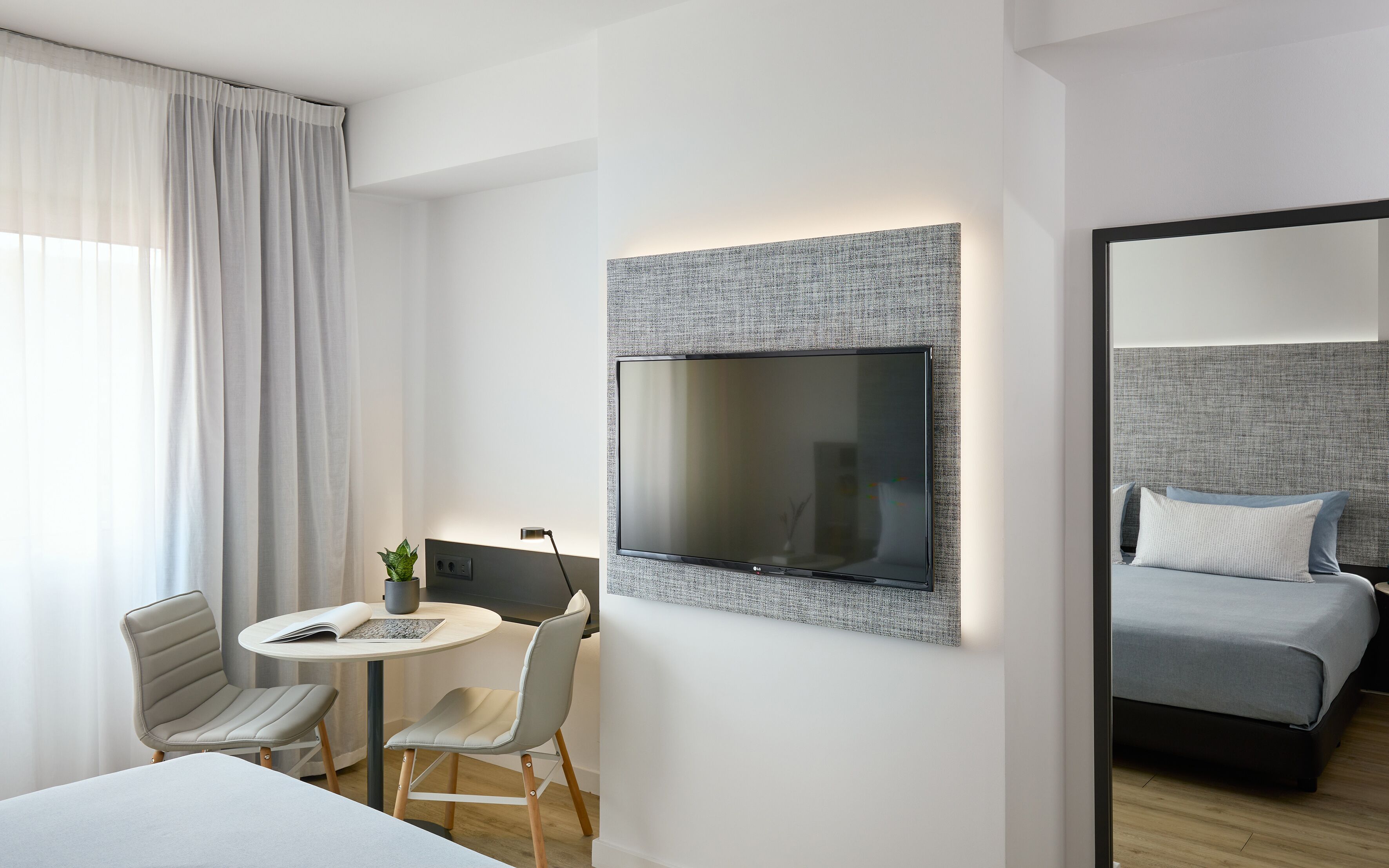 A modern hotel room with a flat-screen TV mounted on a textured wall, a small round table with a chair, a bed with grey bedding, and a large window with sheer curtains.