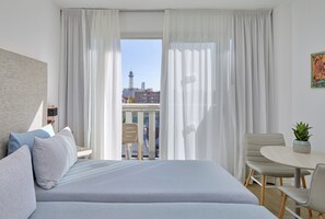 Une chambre d’hôtel avec un lit, un bureau, une chaise et une vue sur le paysage urbain, visible à travers de fines rideaux.