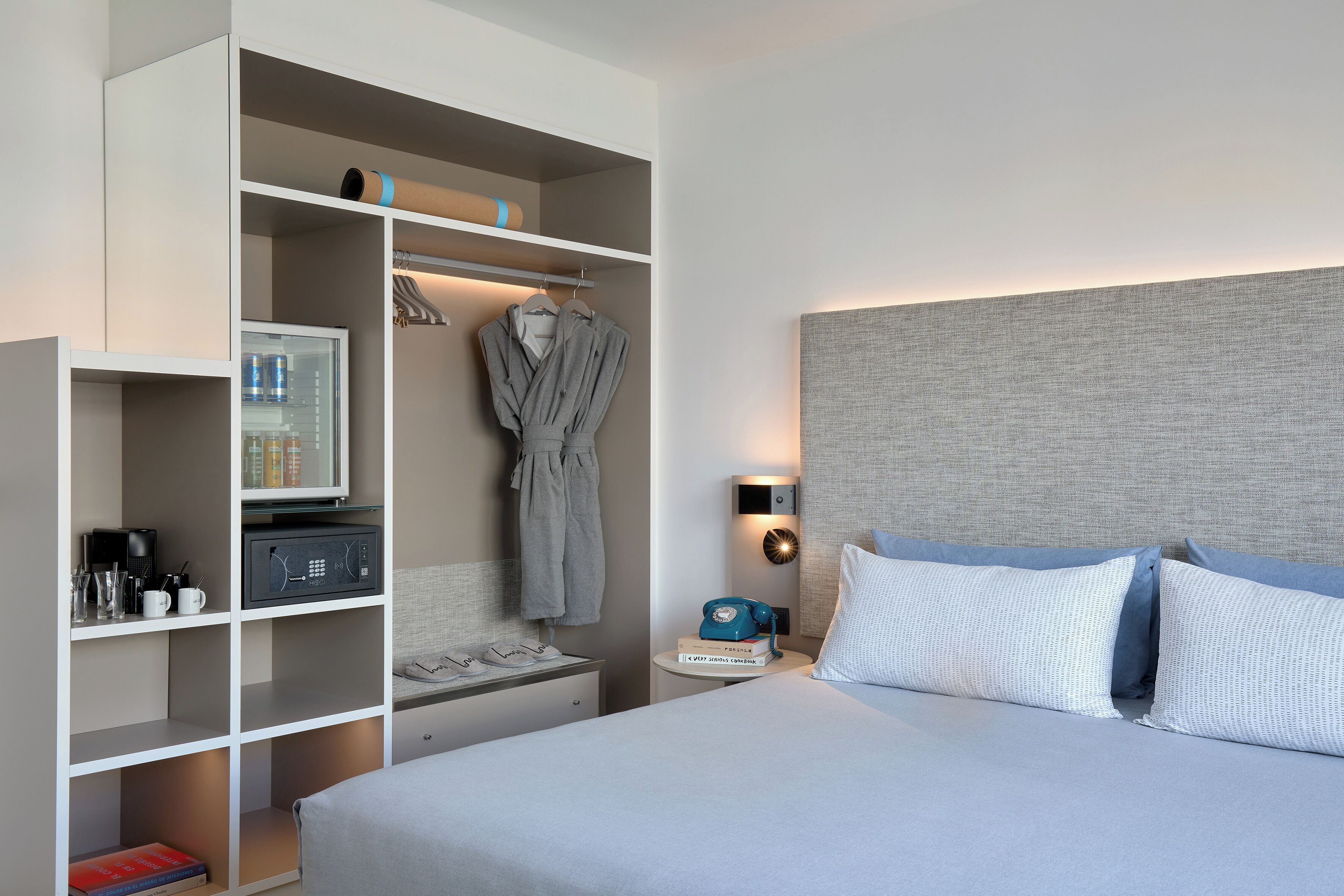 A hotel room with a bed, a wardrobe, a nightstand with a lamp, a telephone, and a shelf with toiletries.