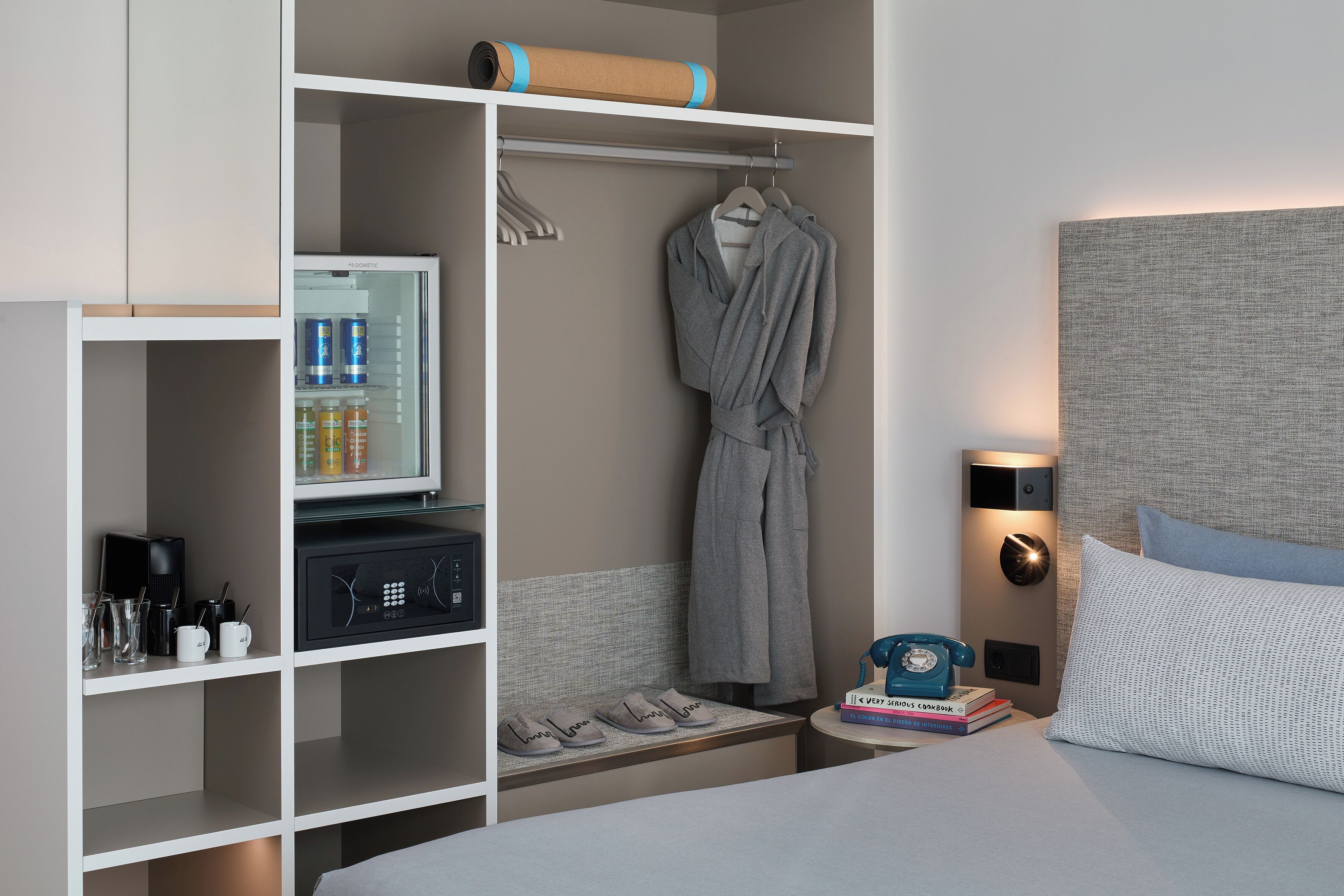 A hotel room with a bed, a bedside table with a telephone and books, a wardrobe with hanging clothes, a shelf with toiletries, and a small seating area with slippers.