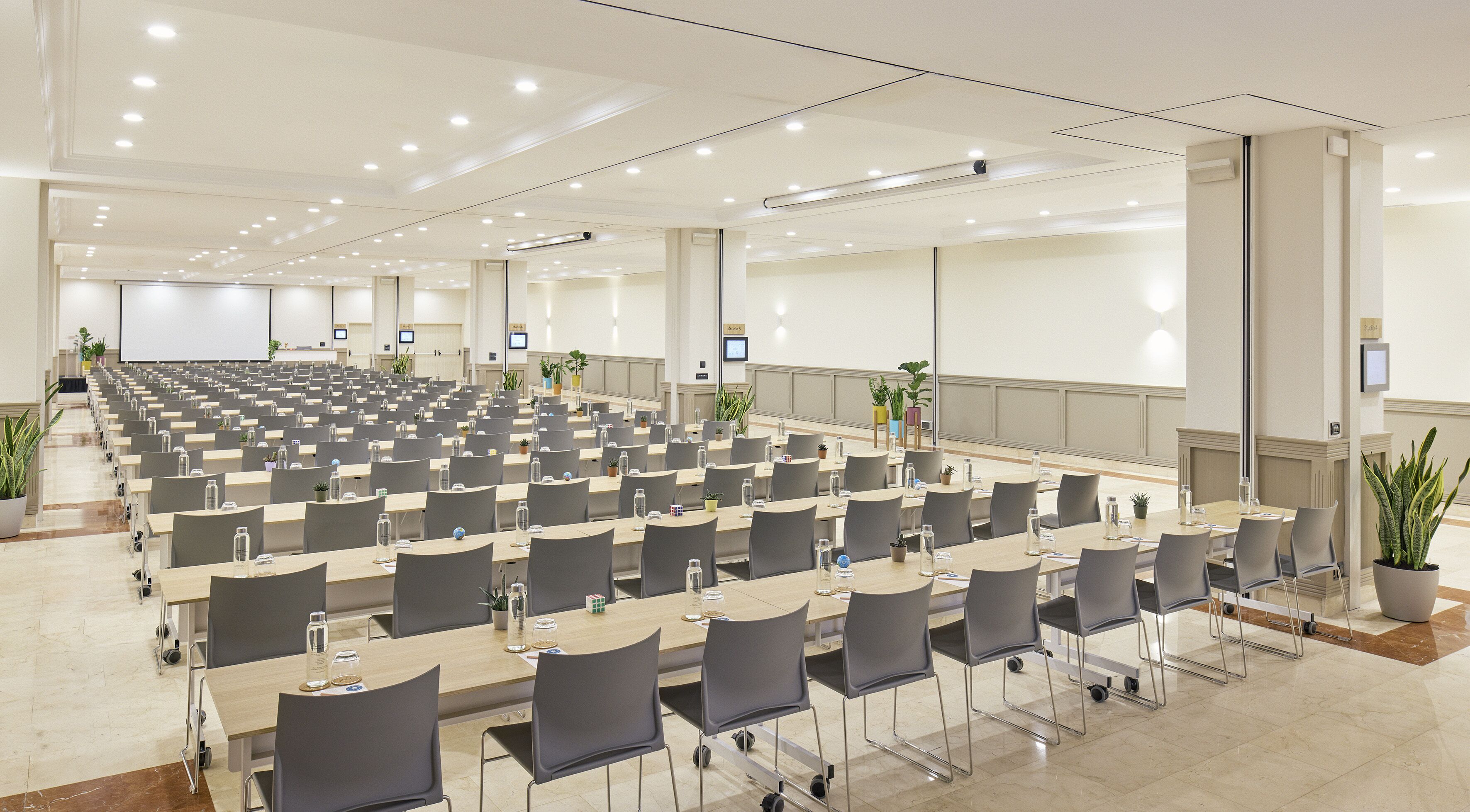 A large conference room with rows of tables and chairs, equipped with water bottles and nameplates, and a large screen at the front.