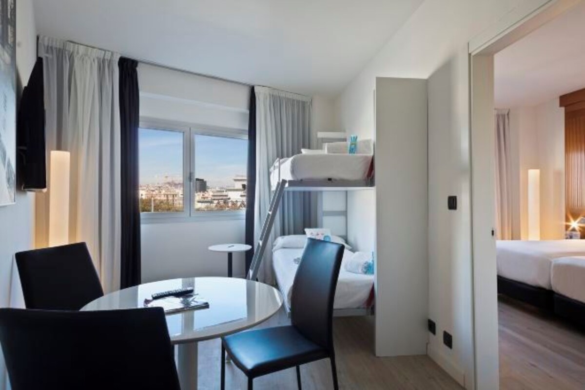 Habitación de hotel con una litera, una mesa redonda de comedor con sillas, un televisor montado en la pared y una ventana con cortinas.