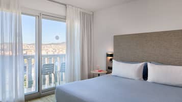 Habitación de hotel con una cama grande, vistas a través de una puerta corrediza de vidrio, una mesita con un florero y vista panorámica de la ciudad.
