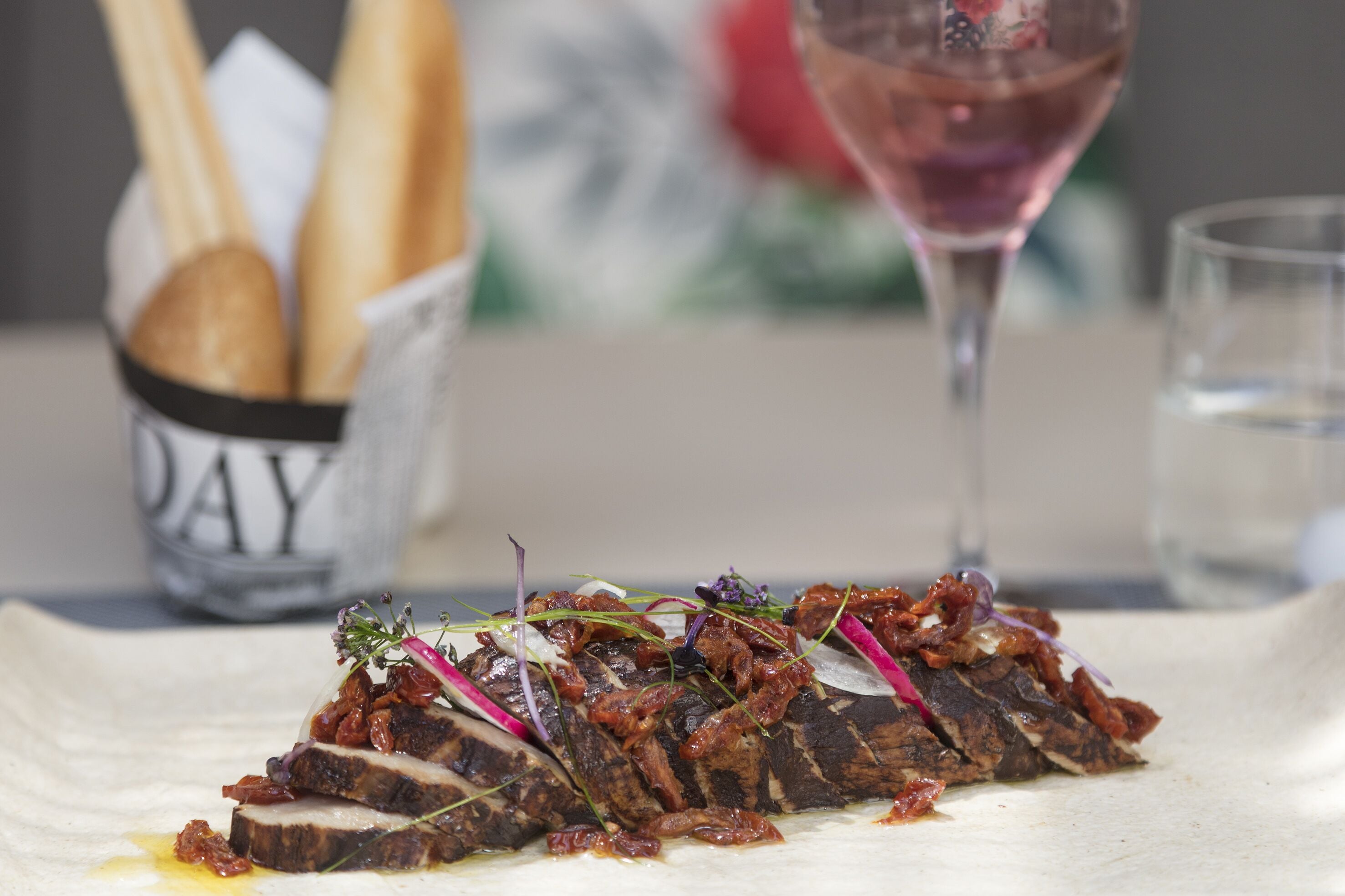 A dish of sliced meat with vegetables and sauce, accompanied by bread and a glass of wine on a table.