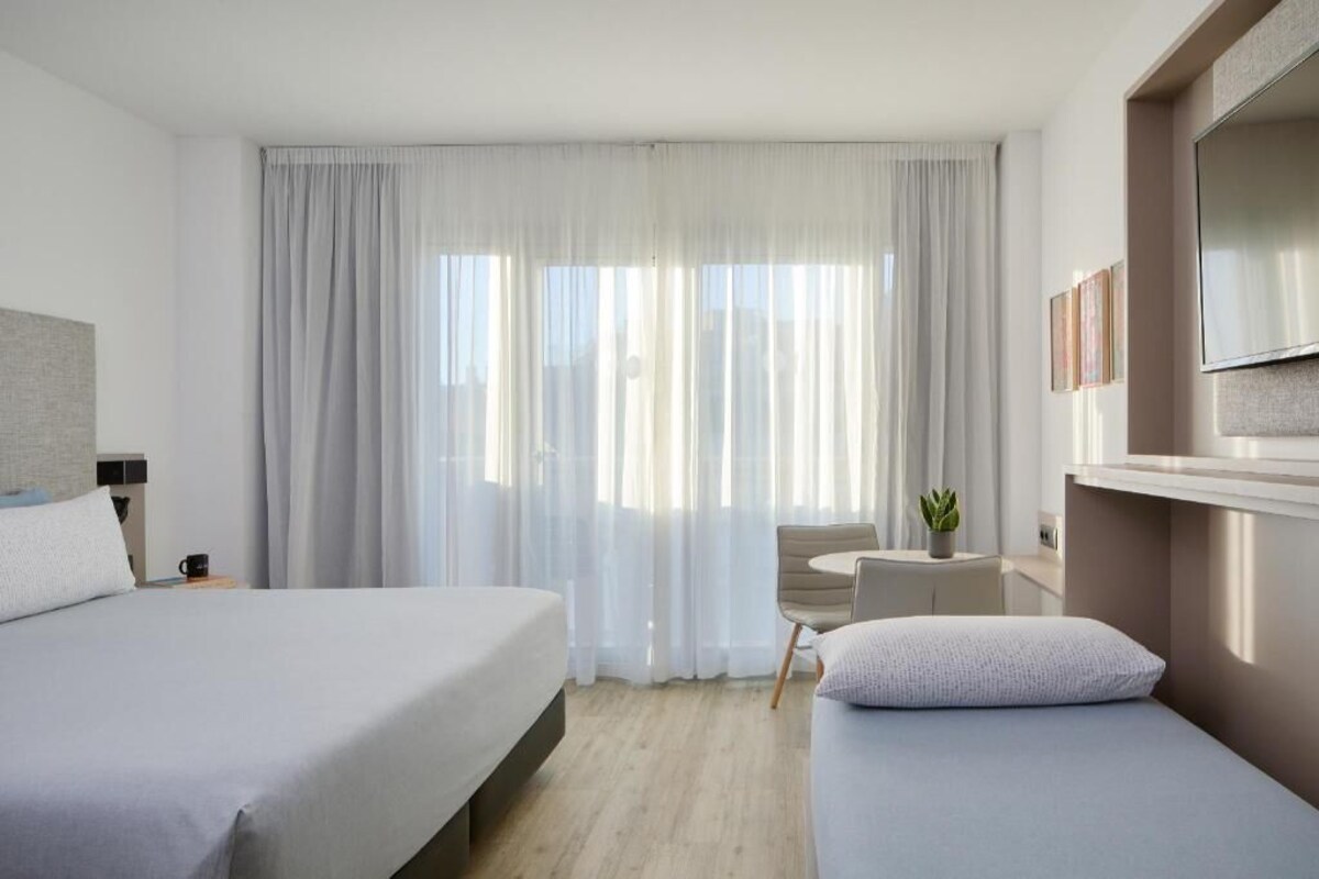 Habitación de hotel con dos camas, televisión, una mesita con una silla y ventana con cortinas translúcidas.