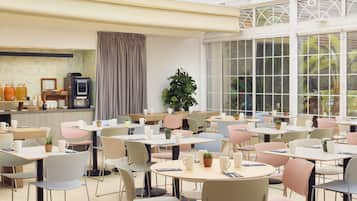 A spacious cafe with multiple tables and chairs, a coffee machine, and a large window with curtains.