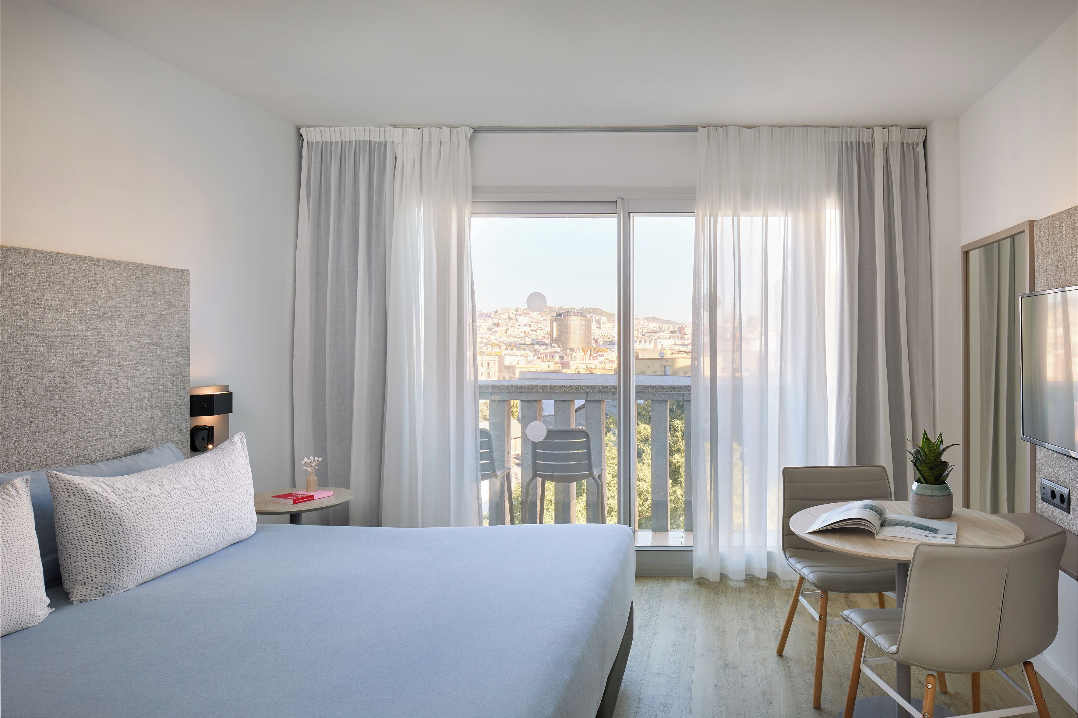A hotel room with a bed, a desk, a chair, a potted plant, and a view of a cityscape through large windows.