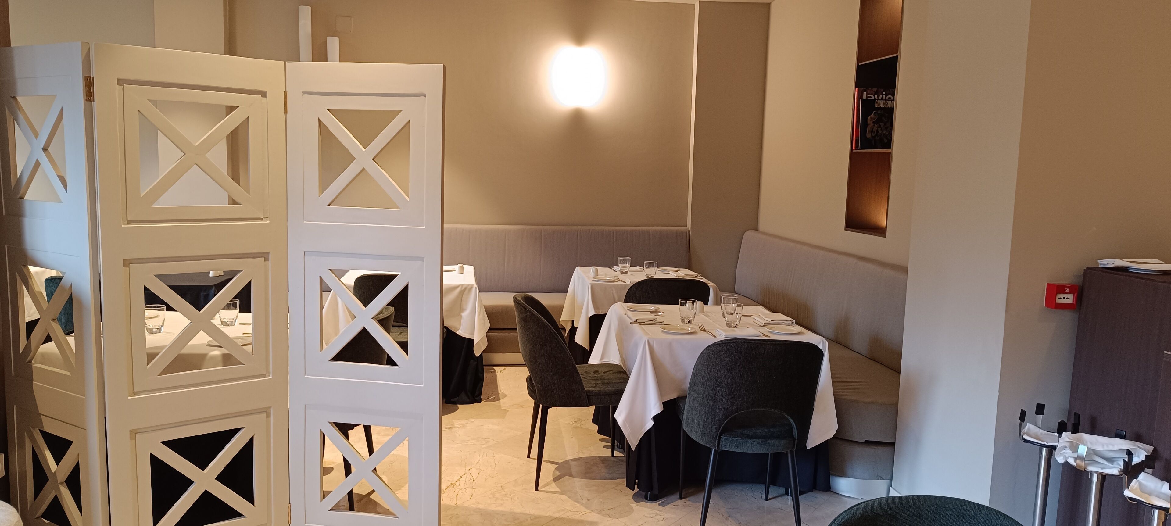 A restaurant with tables set for dining, a light fixture on the wall, and a partition with cut-out designs.