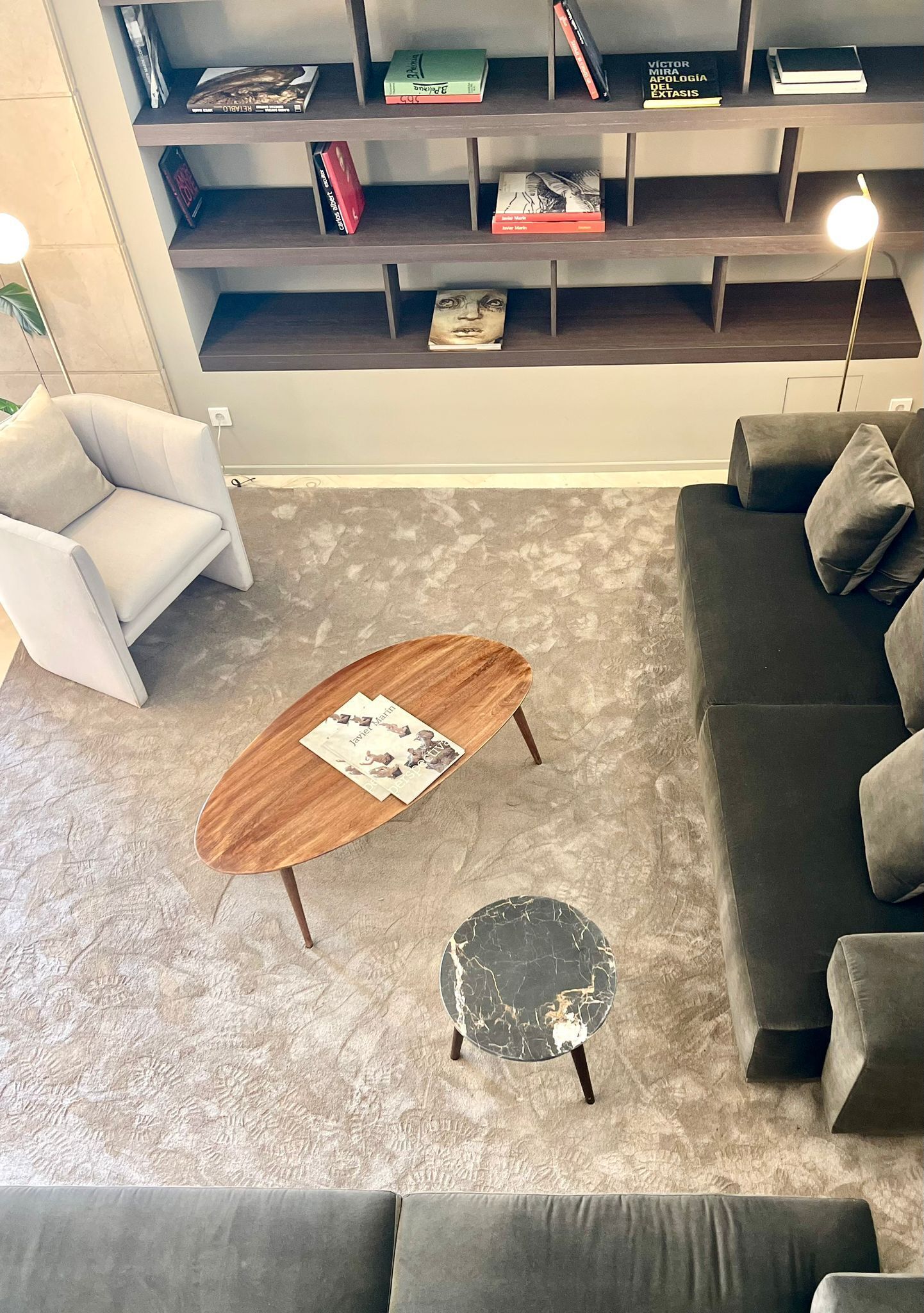 Una sala moderna con un sofá gris, un sillón blanco, una mesa de centro de madera y una mesita auxiliar de mármol negro. Se ve una estantería llena de libros y objetos decorativos al fondo.