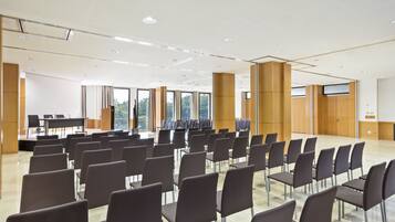 Un salón de conferencias con filas de sillas, un escenario y amplias ventanas.