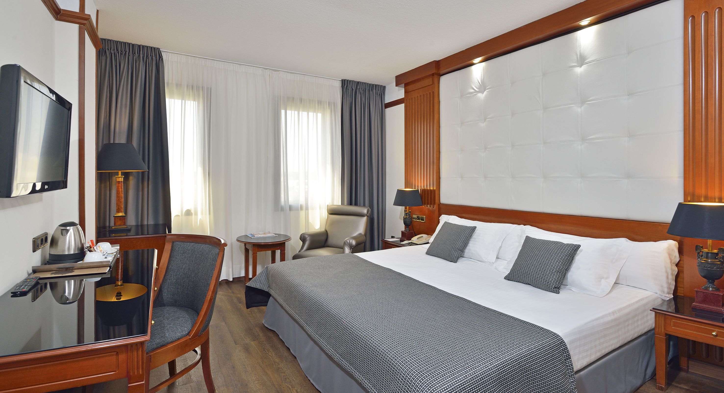 A hotel room with a large bed, a desk with a computer, a chair, a TV, and a bedside table with a lamp.