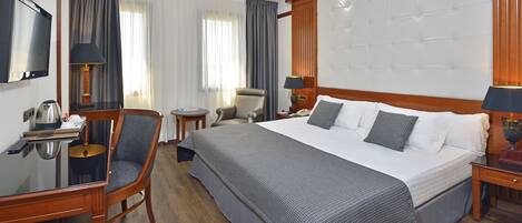 Habitación de hotel con una cama grande, un escritorio con computadora, una silla, un televisor y una mesita de noche con lámpara.