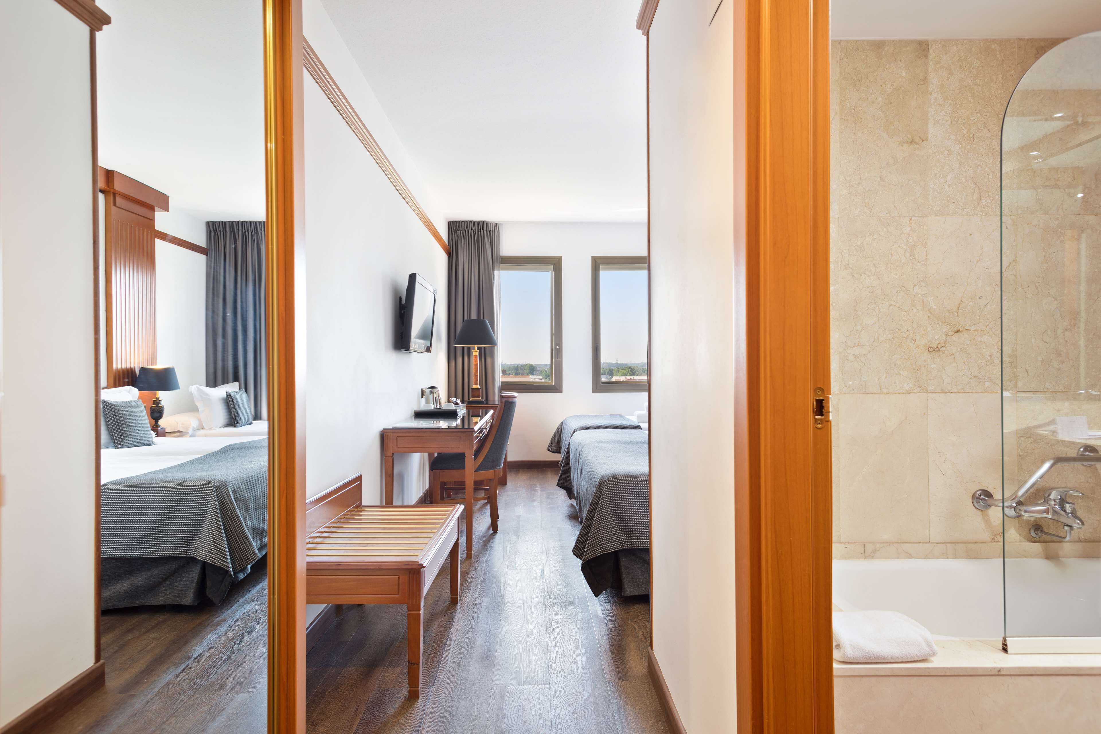 A hotel room with a bed, a desk, a chair, a small table, a TV, and a shower area.