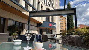 An outdoor seating area with a glass table, a wine glass, and a cup, in front of a building with a sign reading "BWP Premier". - Best Western Premier CMC Girona (Girona)