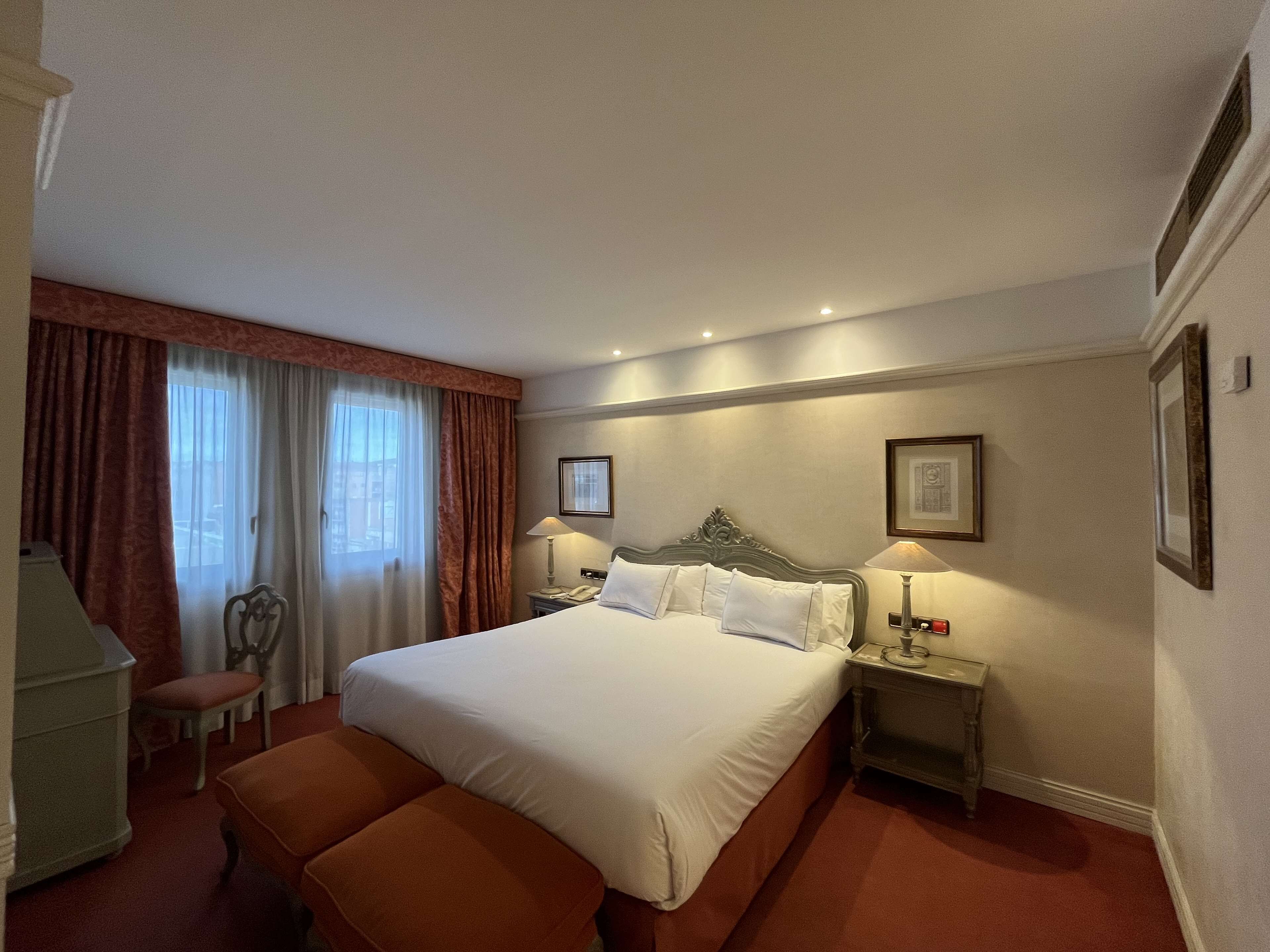 A hotel room with a large bed, two bedside tables with lamps, a chair, a desk, and a window with curtains.