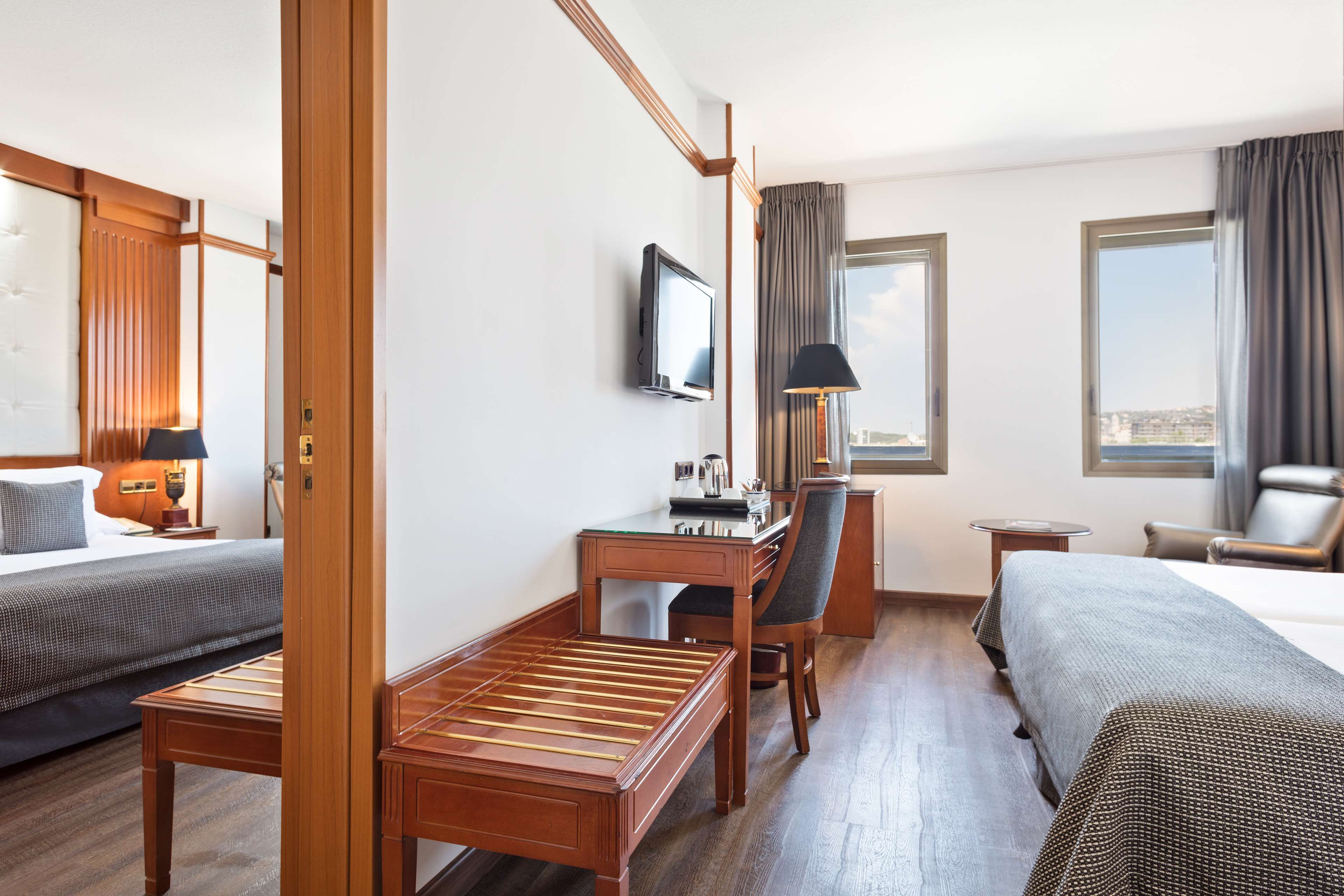 A hotel room with two beds, a desk, a chair, a TV, and a window with curtains.