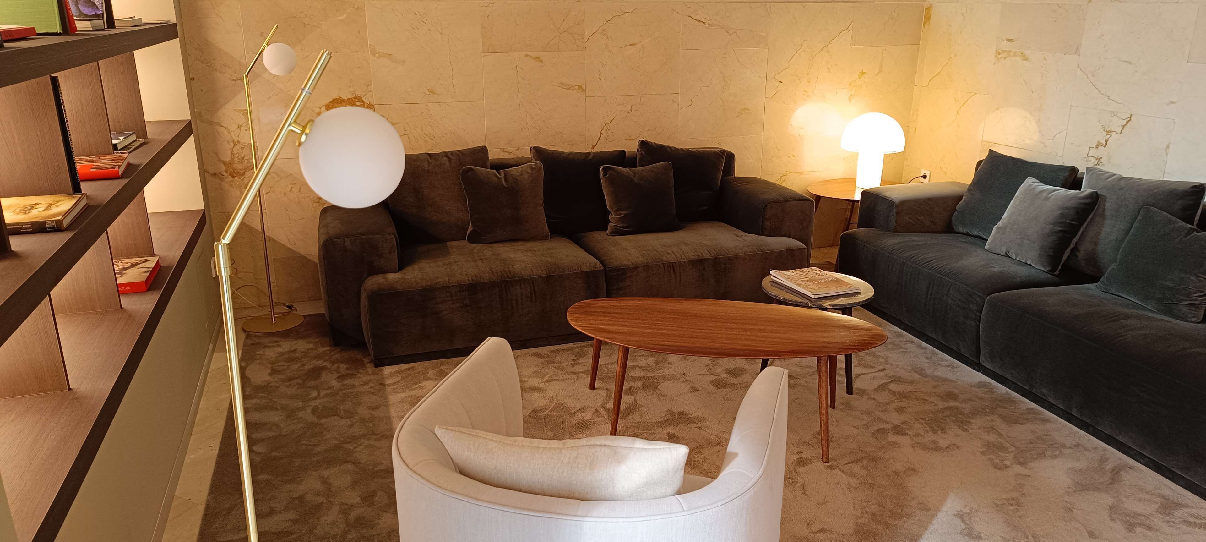 A modern living room with a dark brown sofa, a white armchair, and a wooden coffee table. There are books on the shelves and a floor lamp.