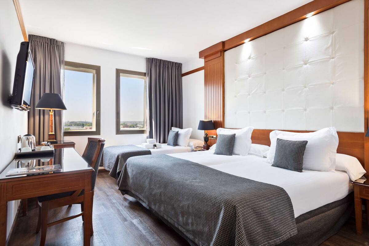 A hotel room with two beds, a desk, a chair, a TV, and a large window.