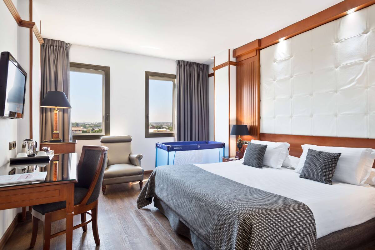 A hotel room with a large bed, a desk with a chair, a television, and a view of the city through the windows.