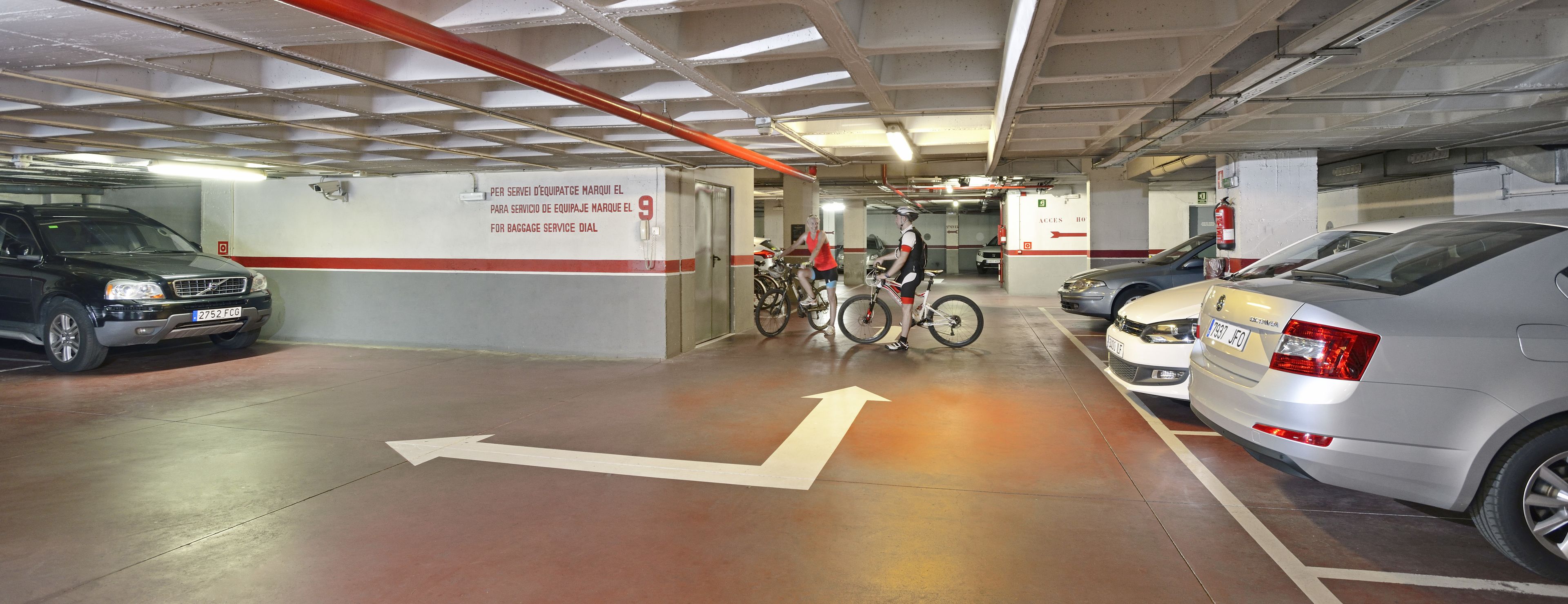 A parking garage with cars and bicycles, directional arrows on the floor, and a wall sign indicating a baggage service area.