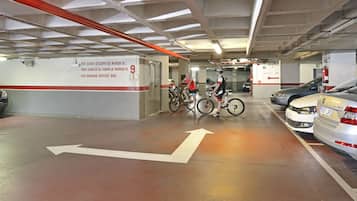 A parking garage with cars and bicycles, directional arrows on the floor, and a wall sign indicating a baggage service area.