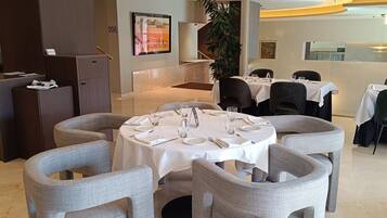 A restaurant with round tables set for dining, a reception desk, and a large plant in the background.