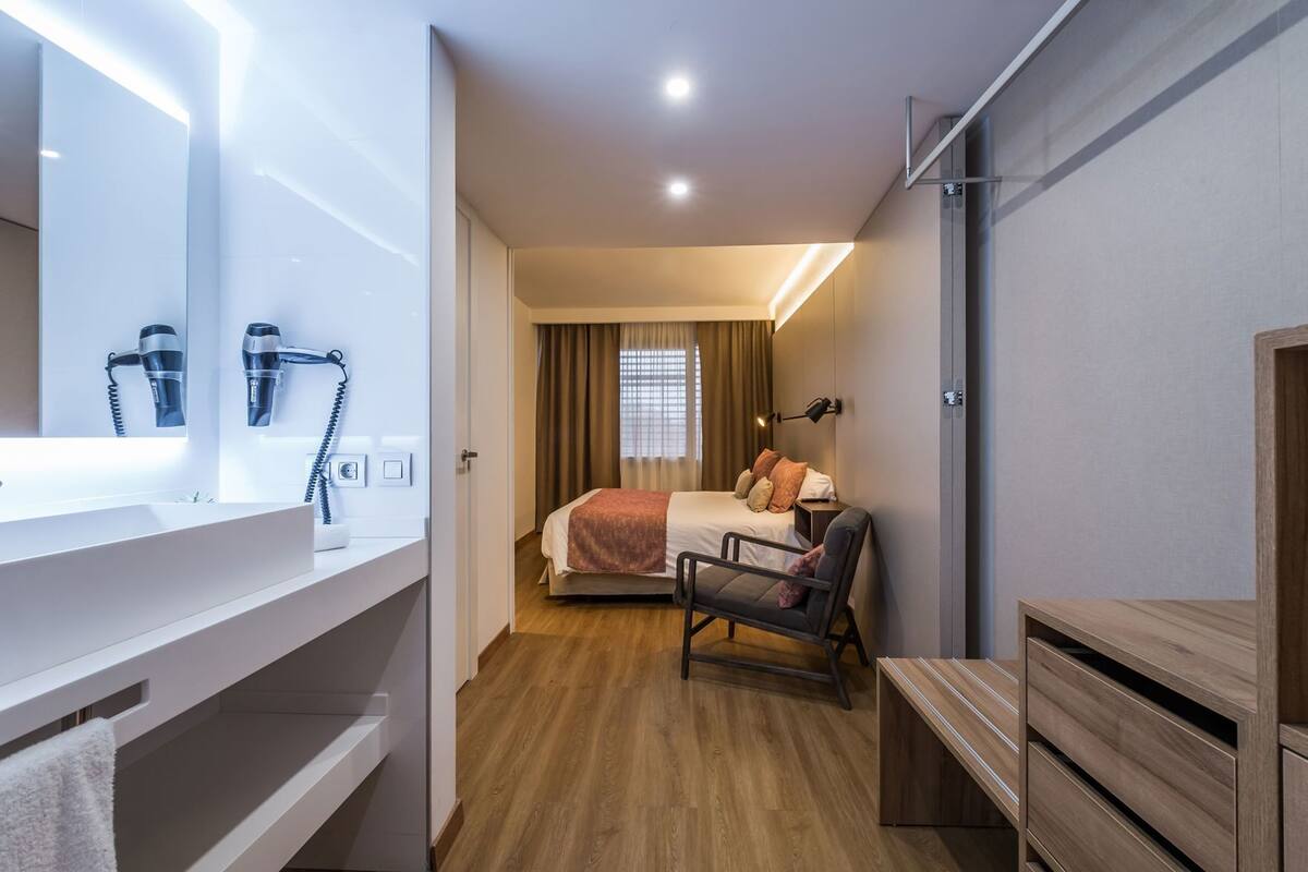 Quarto de hotel com cama, cadeira, escrivaninha, espelho e banheiro com pia e secador de cabelo.