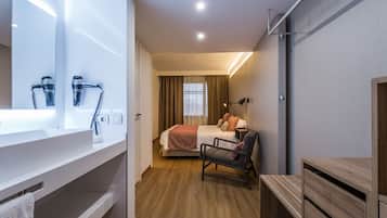 Una habitación de hotel moderna con un espejo grande, piso de madera, cama con una manta color beige, una silla y un armario de madera.