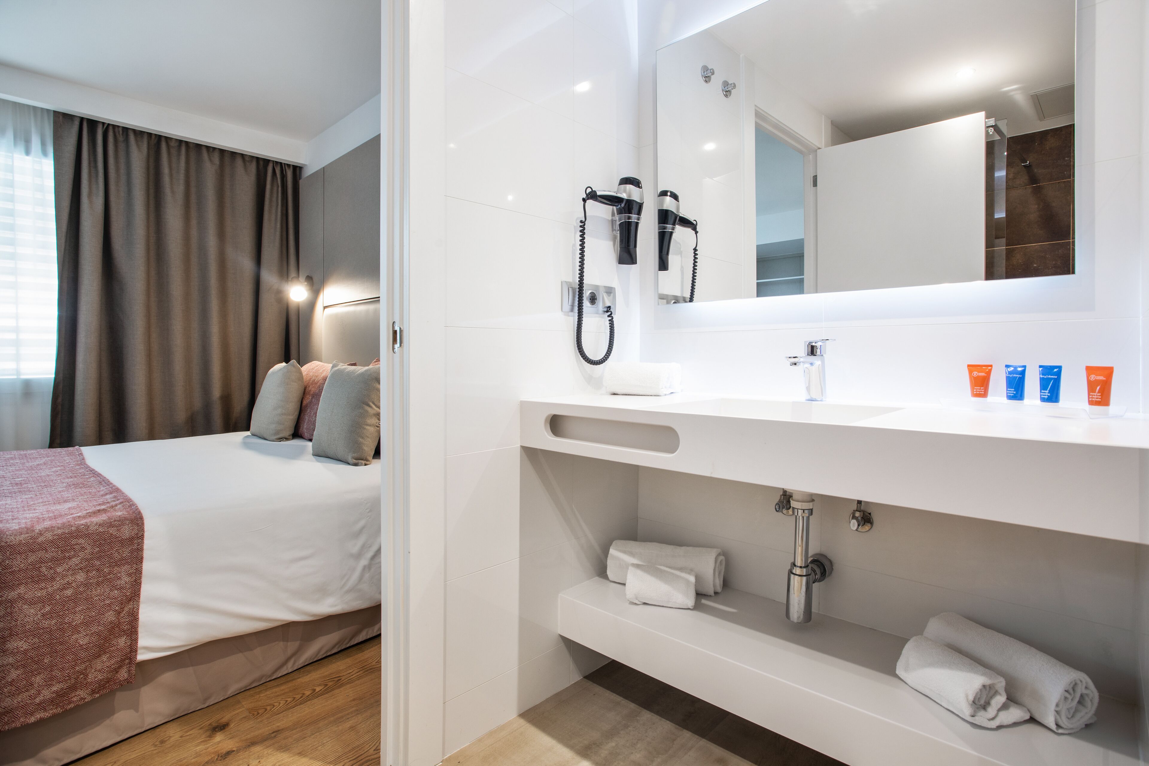 Et moderne hotelværelse med en velopredt seng, et stort spejl, en indbygget toiletbord med vask og et bruseområde.
