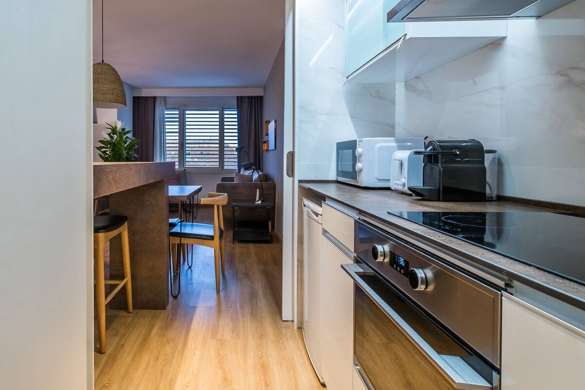 Cozinha moderna com forno embutido, micro-ondas e torradeira. Possui área de jantar com mesa e cadeiras, e uma janela com persianas.