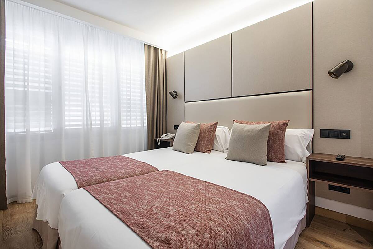 Quarto de hotel com uma cama grande, uma mesa de cabeceira, uma luminária e uma janela com cortinas.