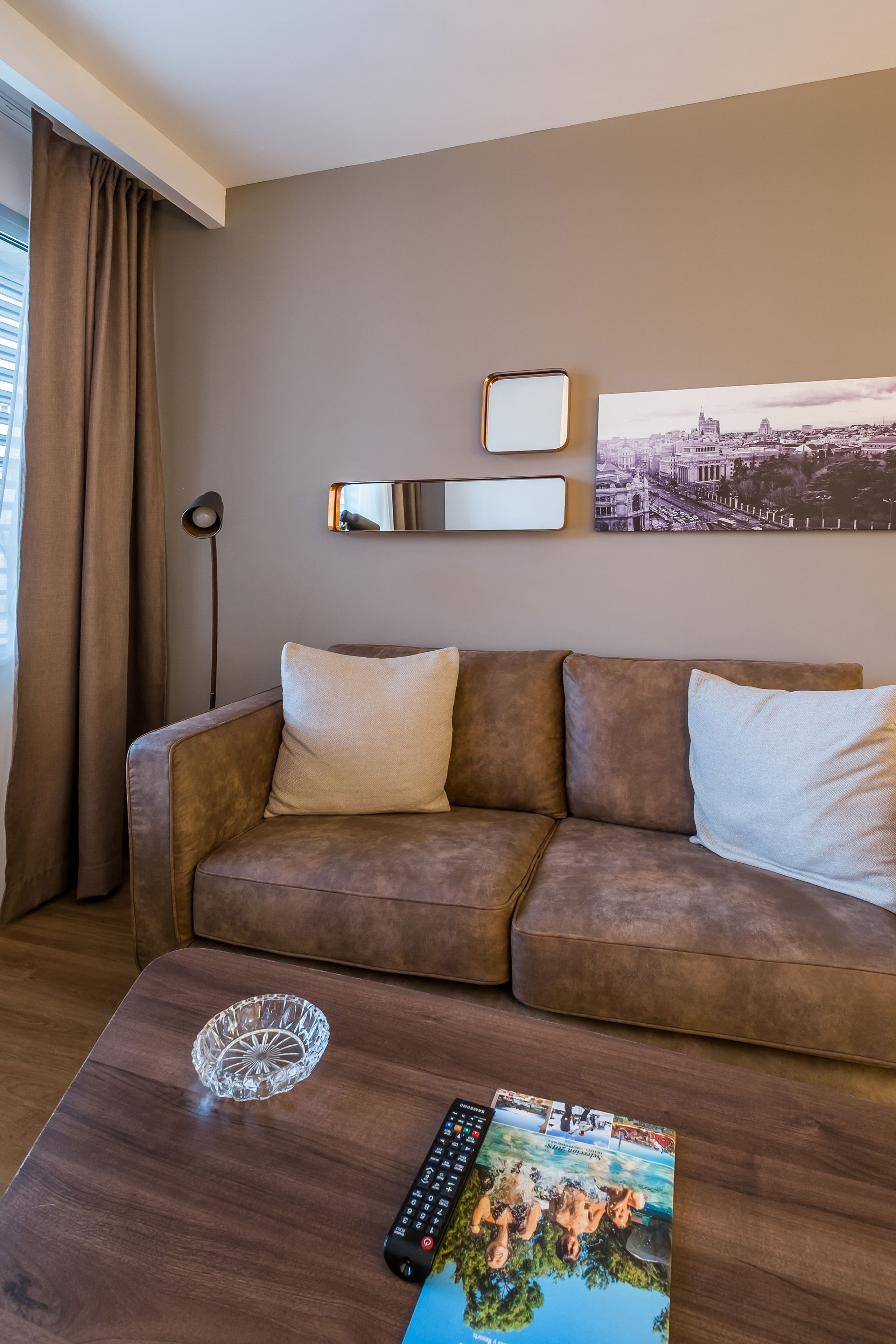 A modern living room with a brown sofa, a glass coffee table, a remote control, and a framed cityscape picture.