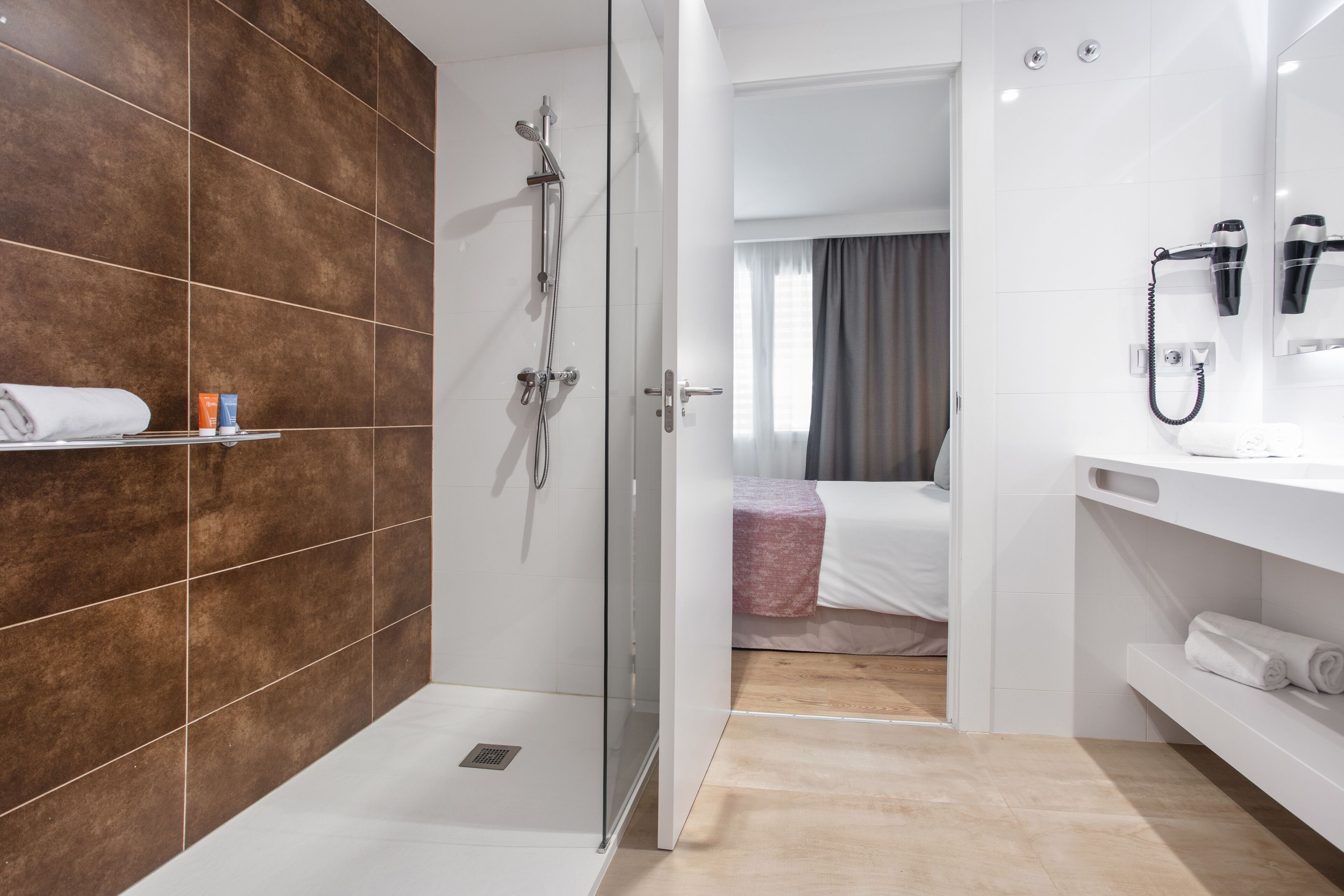 A modern bathroom with a glass-enclosed shower, a white sink with a hairdryer, and a bed with a pink blanket.