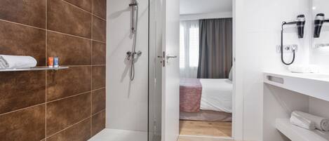 A modern bathroom with a glass-enclosed shower, a white sink with a hairdryer, and a bed with a pink blanket.