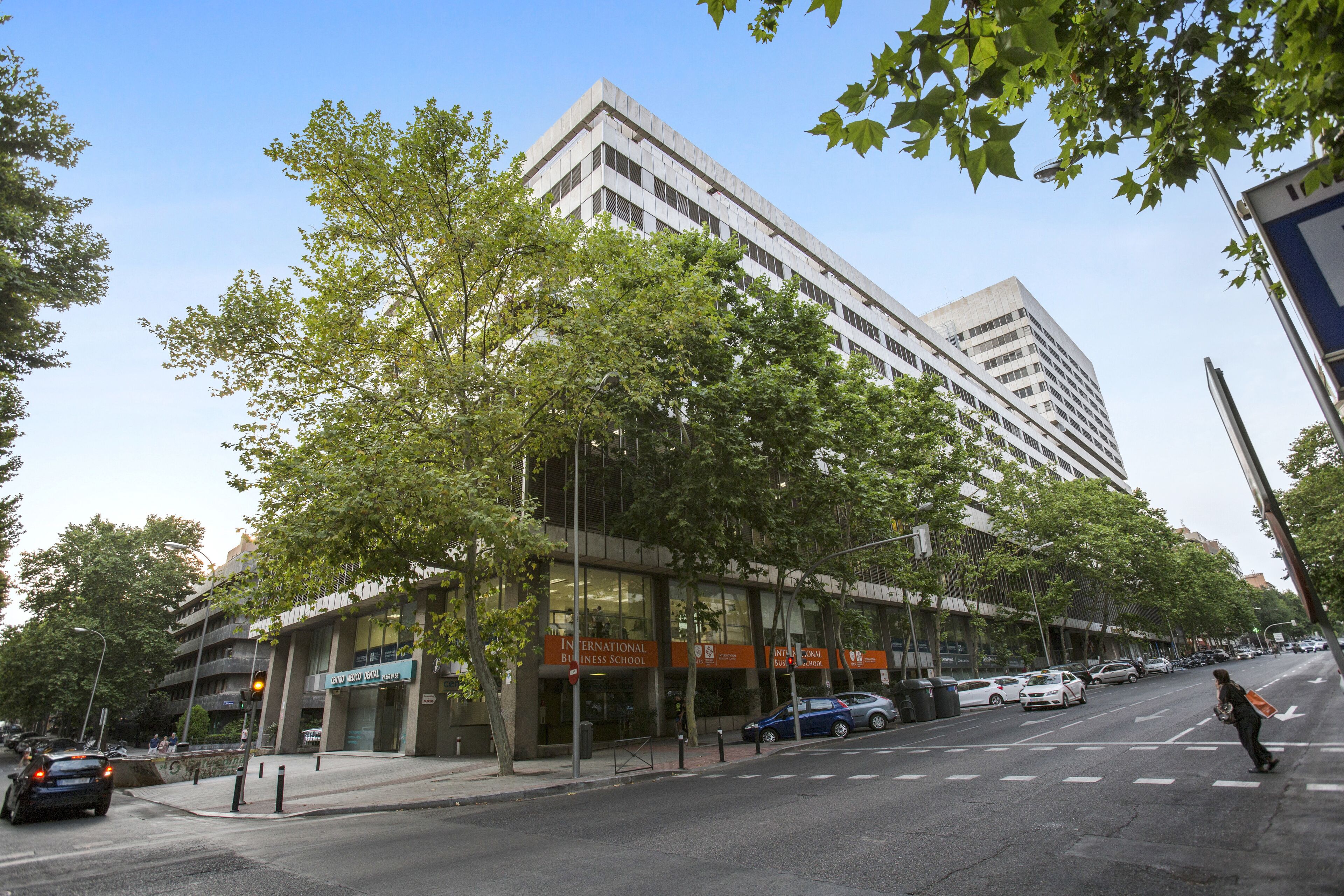 A modern building with a glass facade, a sign for an International Business School, and a street with parked cars and a pedestrian crossing.