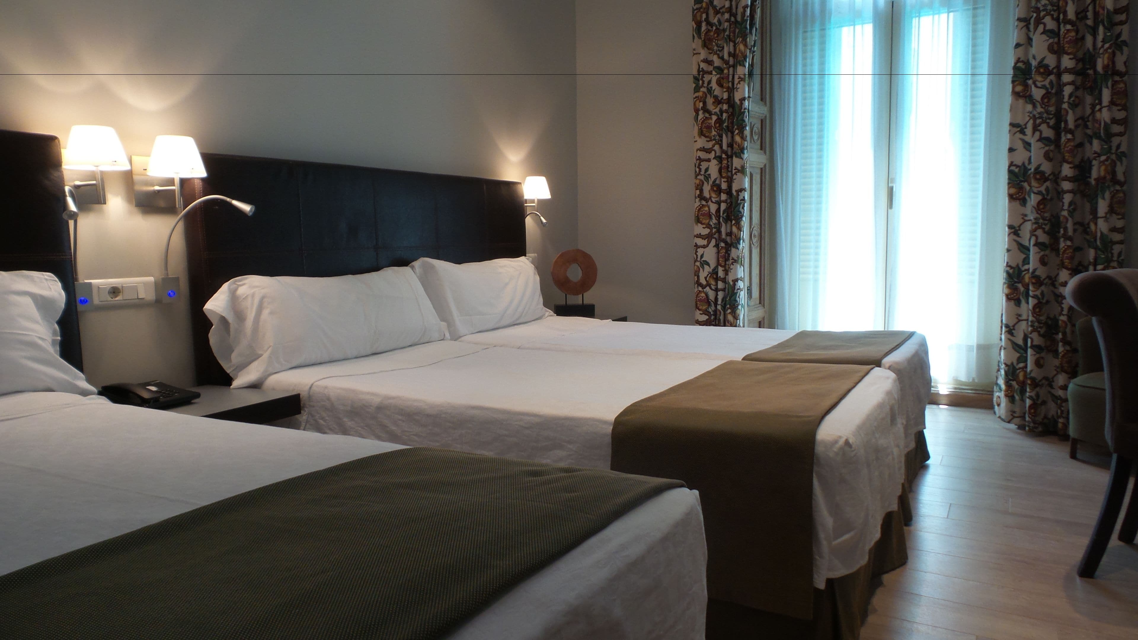 Habitación de hotel con dos camas, una mesita de noche con lámpara, un enchufe y una ventana con cortinas.