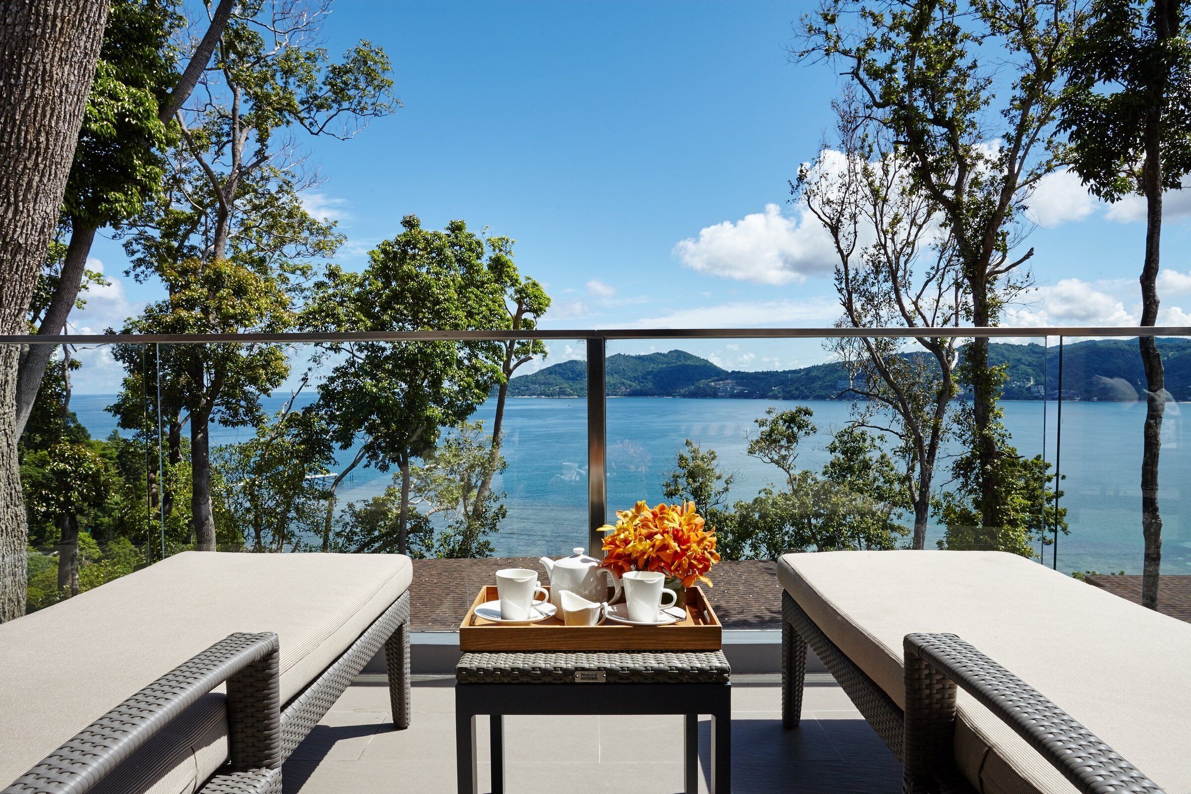 A balcony with a view of a lake and mountains, a tray with cups and a flower, and two cushioned chairs.