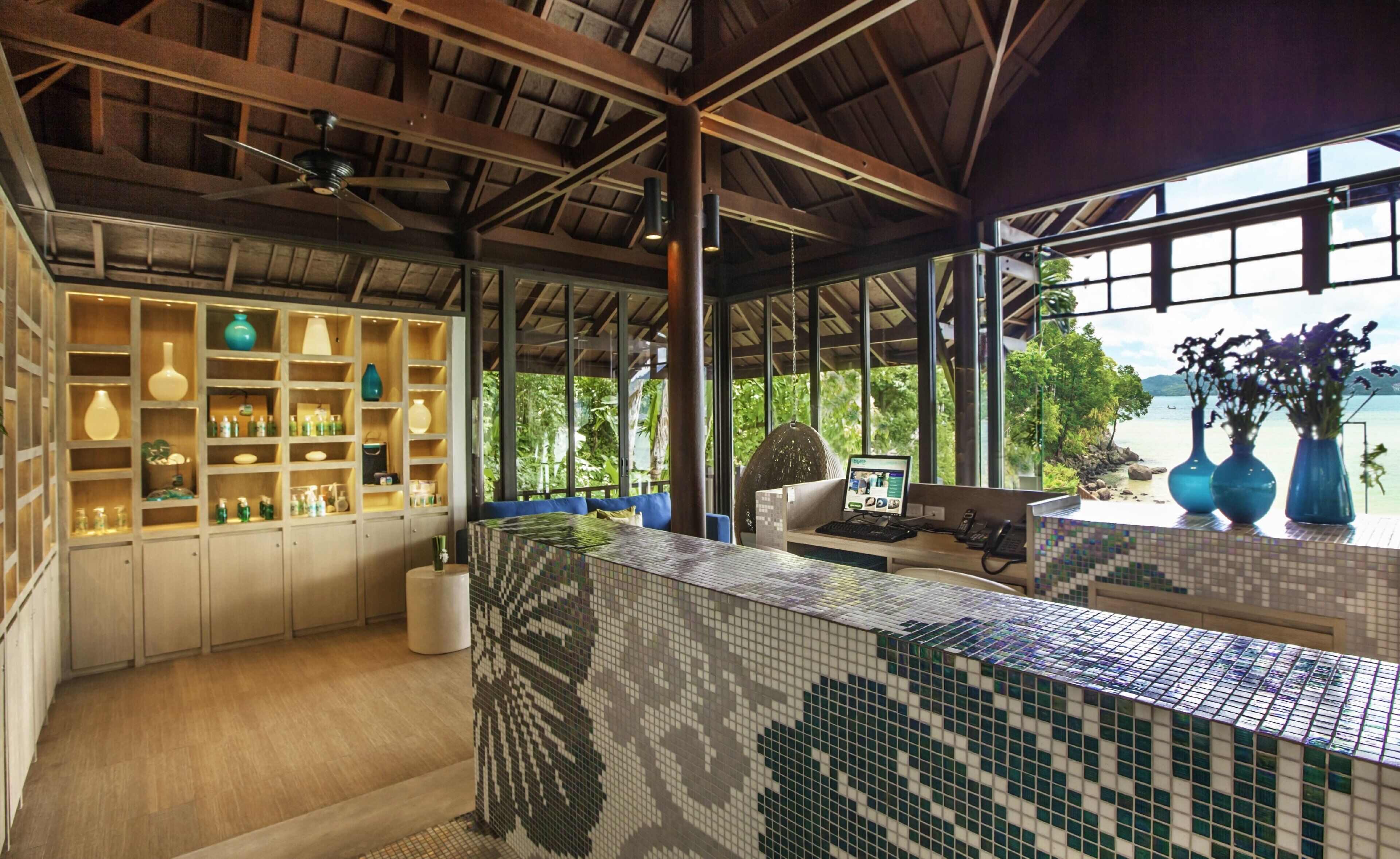 Amplia habitación con salpicadero de azulejos, estantes de madera con diversos objetos, una silla colgante y vista al mar a través de amplias ventanas.