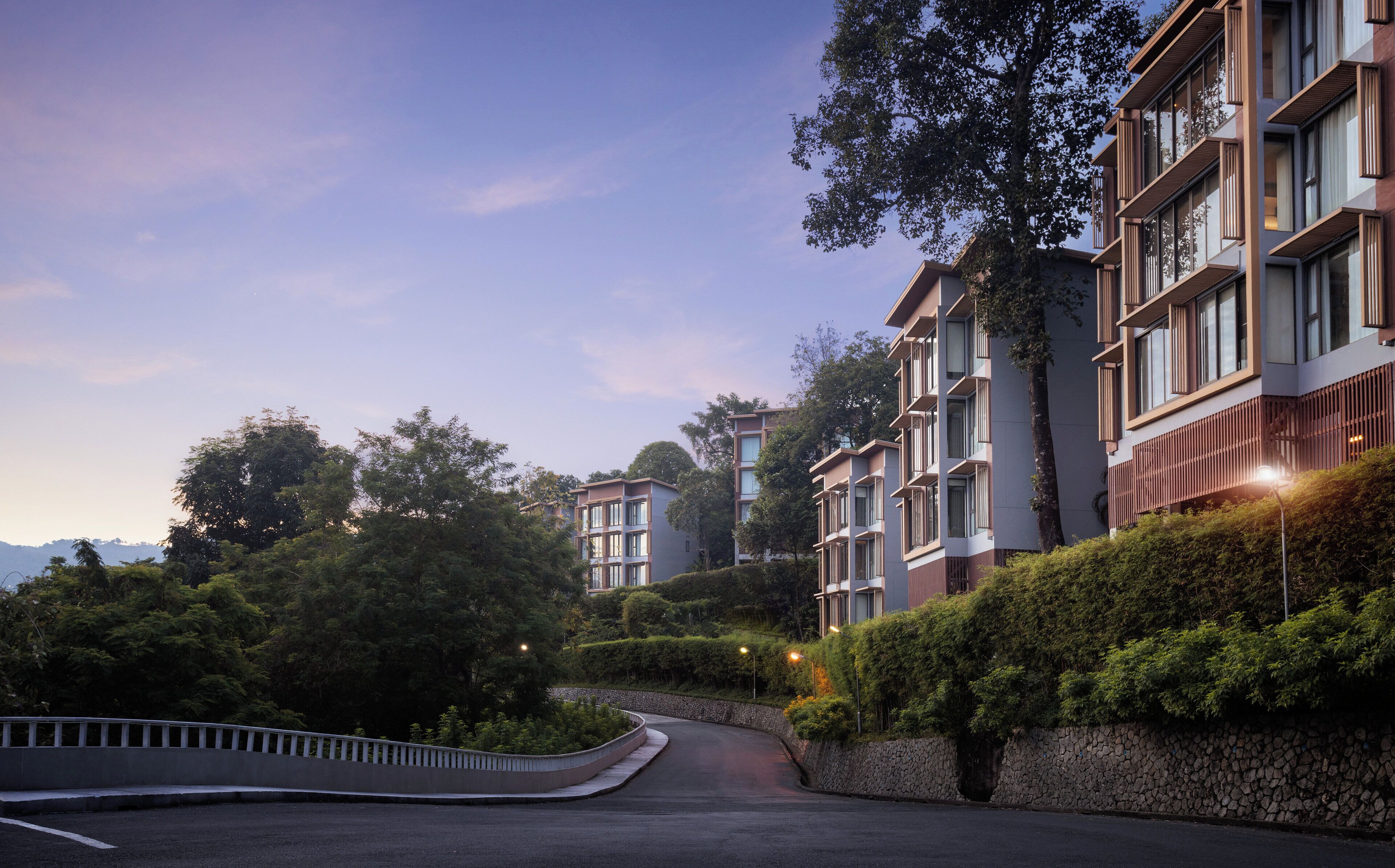 A modern residential building with multiple balconies, surrounded by lush greenery and a curved road.