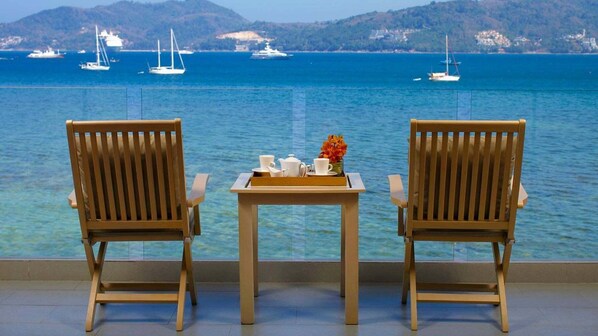 Two wooden chairs facing a table with a tray of tea set and a vase of flowers, overlooking a calm sea with boats and hills in the background.