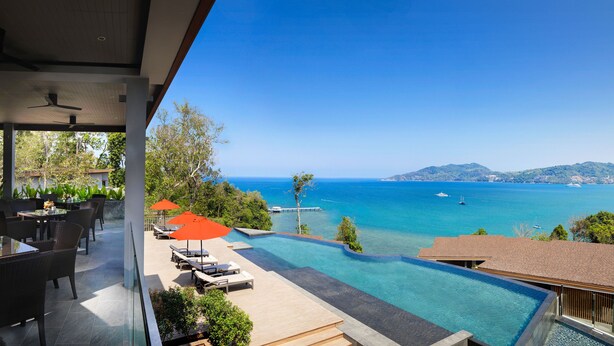 Un área exterior moderna con piscina, sillones de descanso y sombrillas, con vista al mar azul y cristalino.